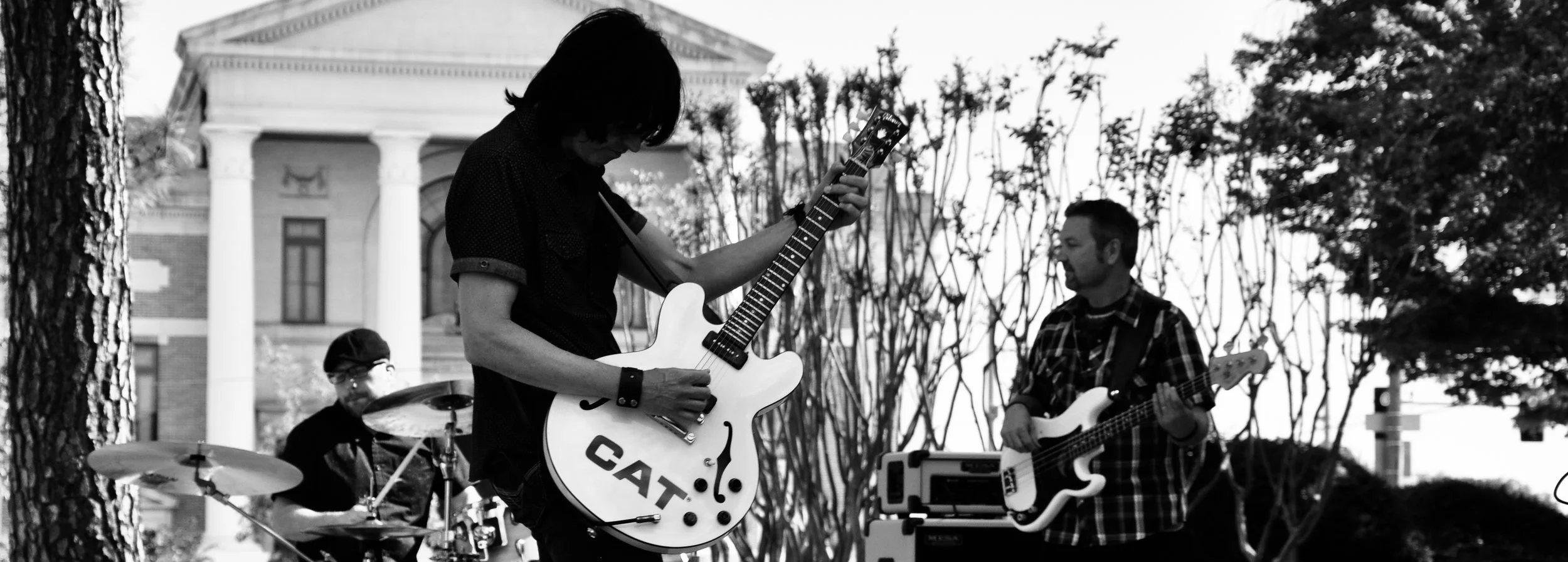 A black and white photo of a band performing outdoors with three musicians. The guitarist with long dark hair in the foreground holds a guitar with the word 'CAT' on it. Behind them, a drummer is playing, and to the right, a bass guitarist is looking at the guitarist. In the background, there is a historic building with columns and trees.