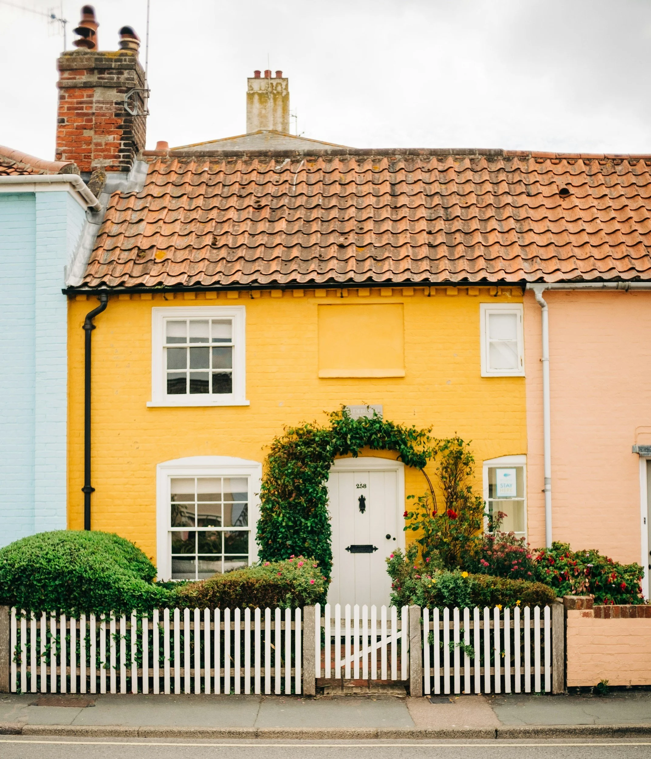 Yellow house with white door and window frames, surrounded by garden with bushes and flowers, white picket fence in front.