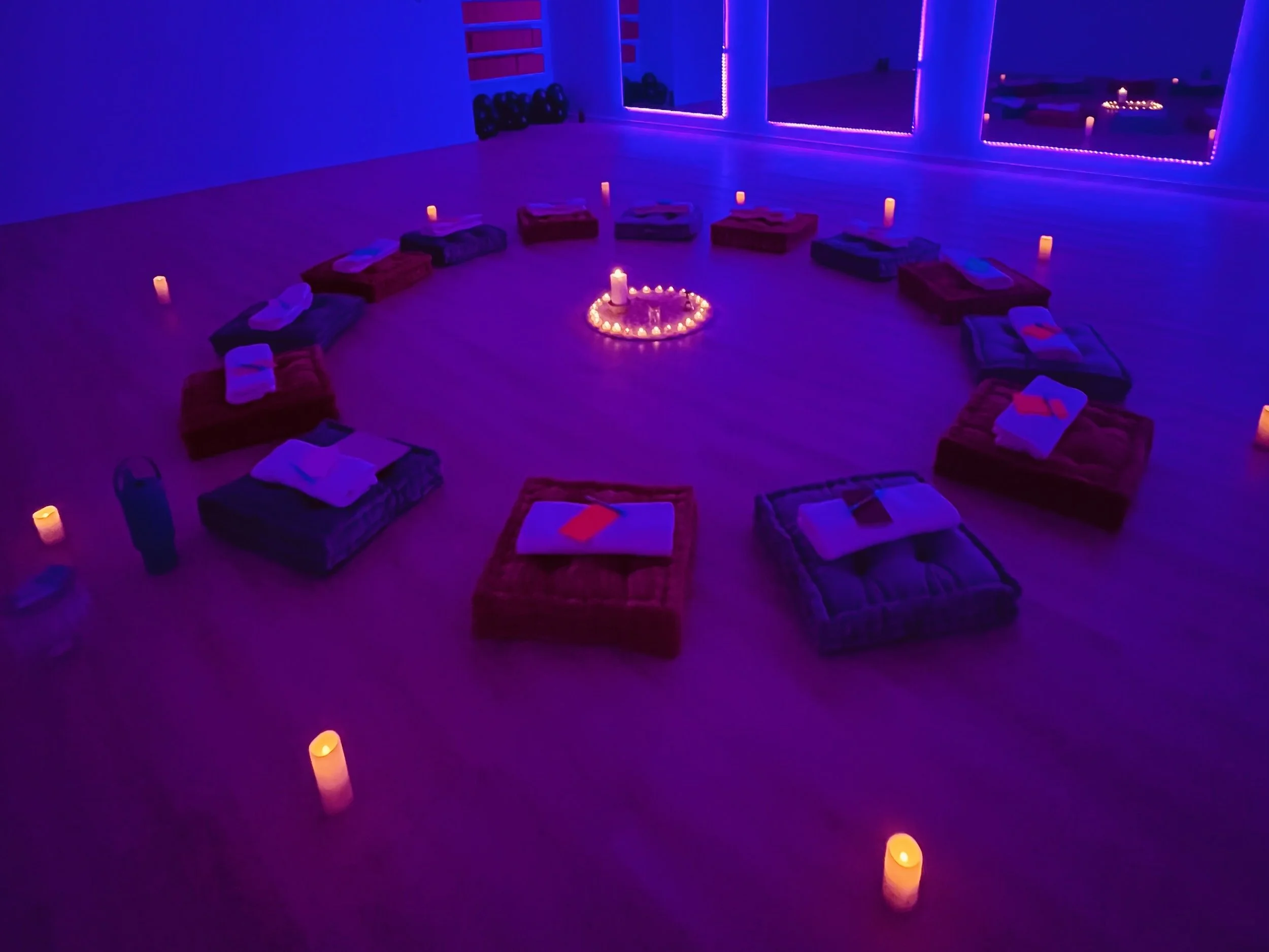 Yoga mats and towels arranged in a circle on the floor, lit by candles, with a candle in the center surrounded by smaller candles, in a dimly lit room with purple and blue lights.