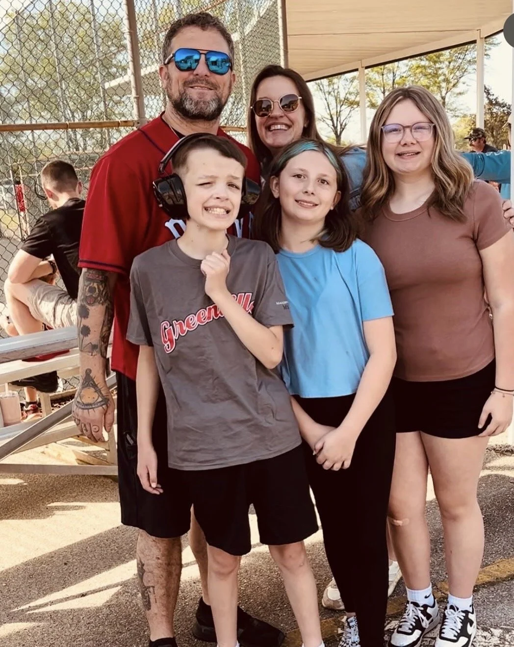 A family group photo outdoors at a park with a chain-link fence, three kids and two adults smiling and posing for the camera.