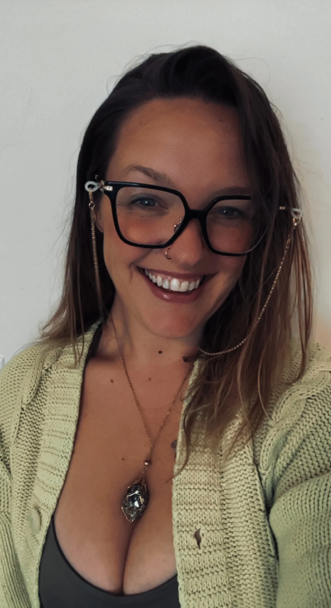 A woman with long hair, glasses with chains, and nose piercings smiling at the camera, wearing a green cardigan and a dark top with a large pendant necklace, against a plain light-colored wall.