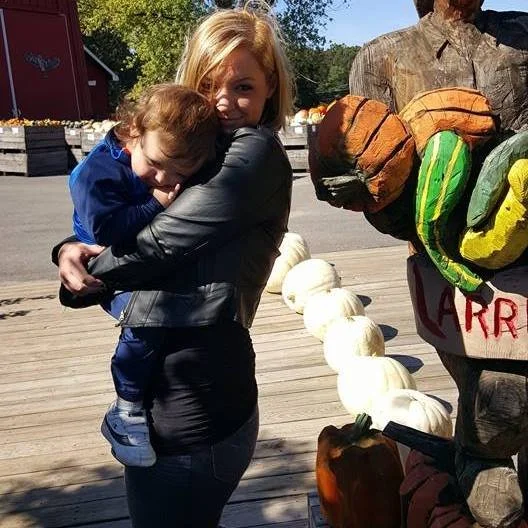 A woman hugging a young boy outdoors, with a pumpkin display and a scarecrow holding painted gourds in the background.