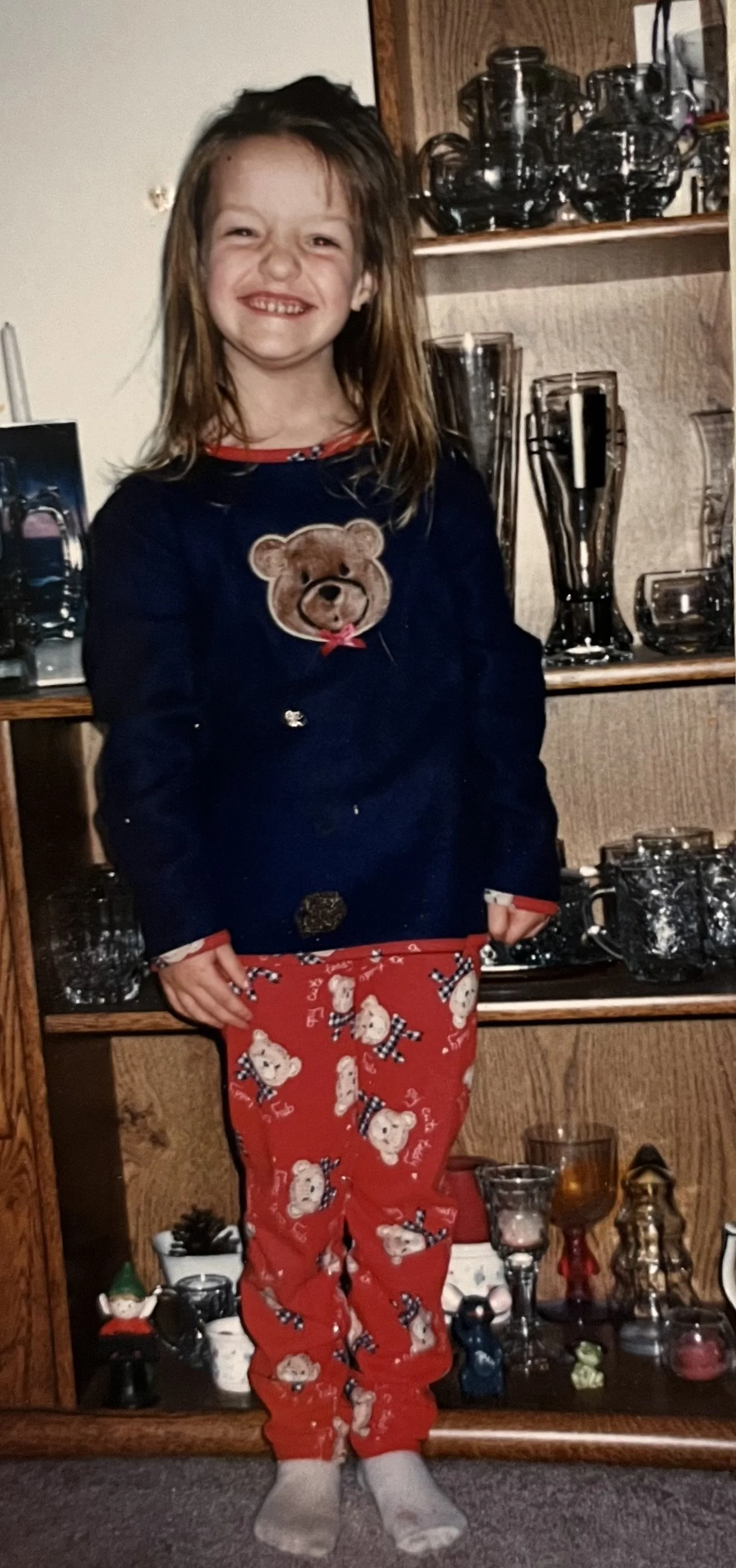 A young girl with long, messy hair standing in front of a wooden display cabinet filled with glassware, wearing red holiday-themed pajamas with teddy bears and a navy blue shirt with a teddy bear graphic.