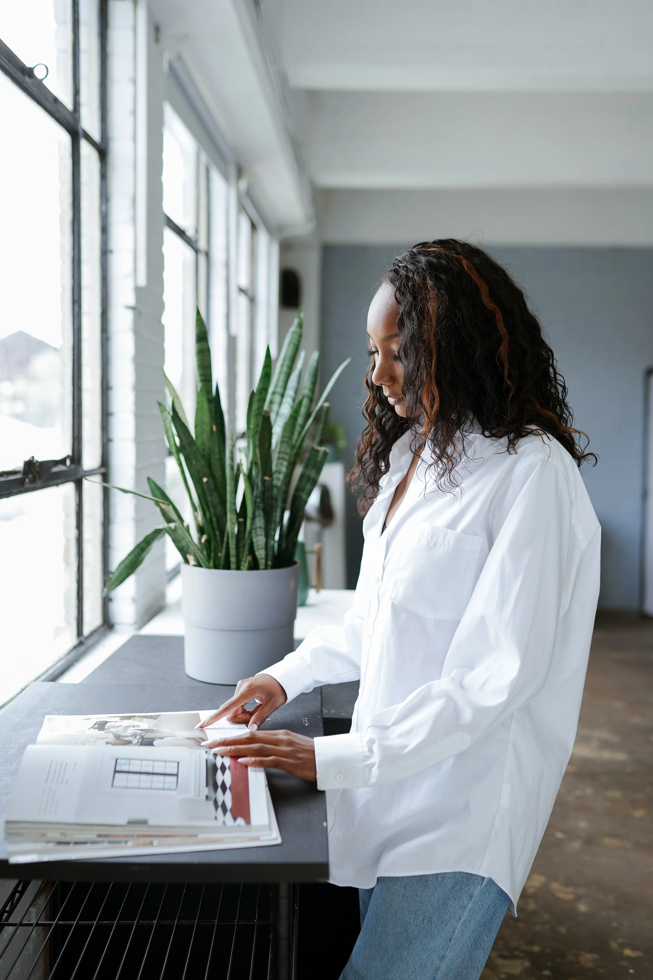 Woman in white shirt looking at a catalog on a black table near a large window, with a tall green plant in a white pot beside her.