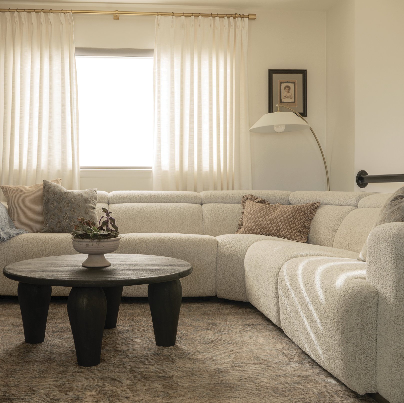 Living room with a beige sectional sofa, decorative pillows, a coffee table with a potted plant, a floor lamp, and curtains covering a window.