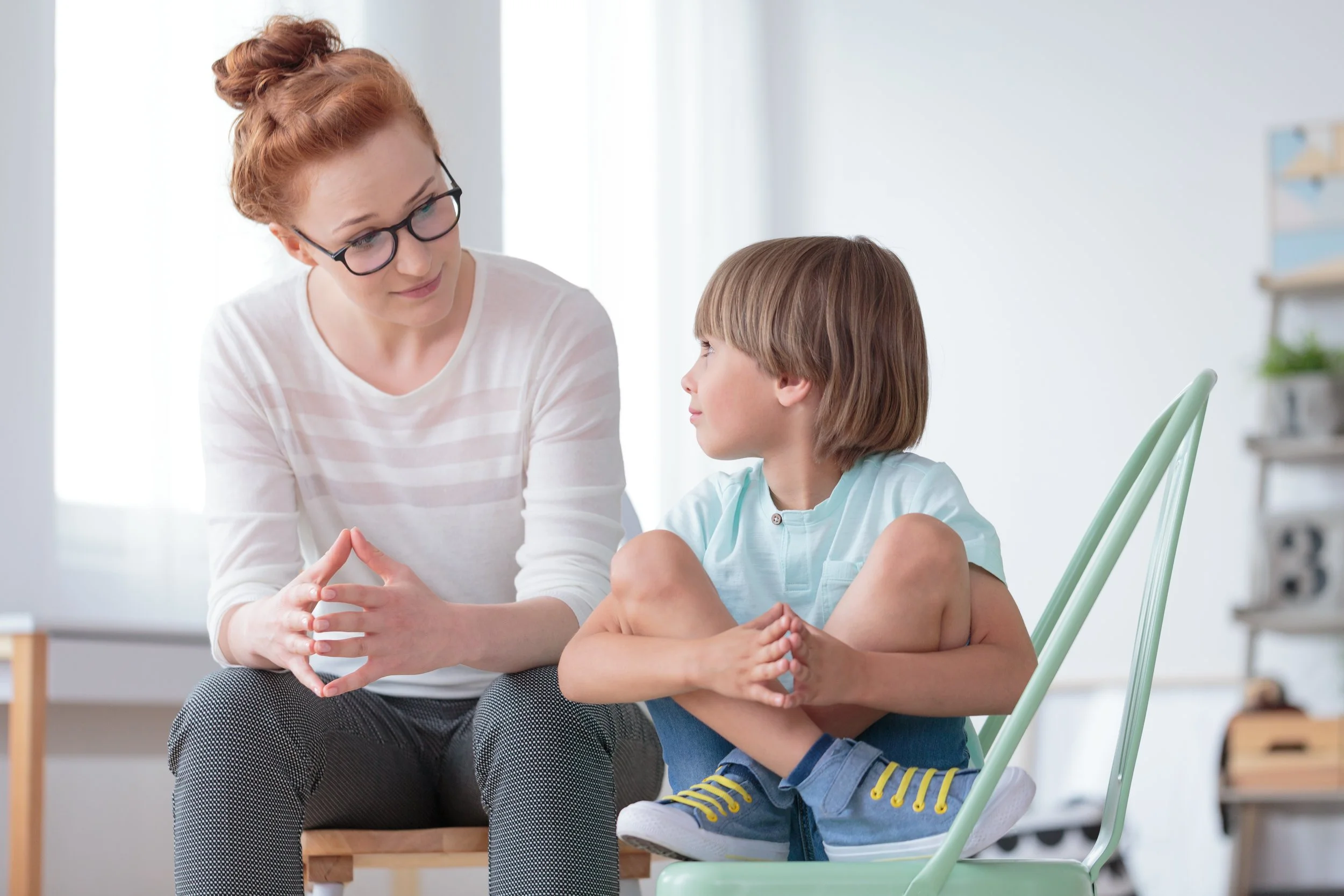 A woman and a young boy sitting indoors, having a conversation, with the woman leaning forward and the boy sitting on a green chair with his legs crossed.