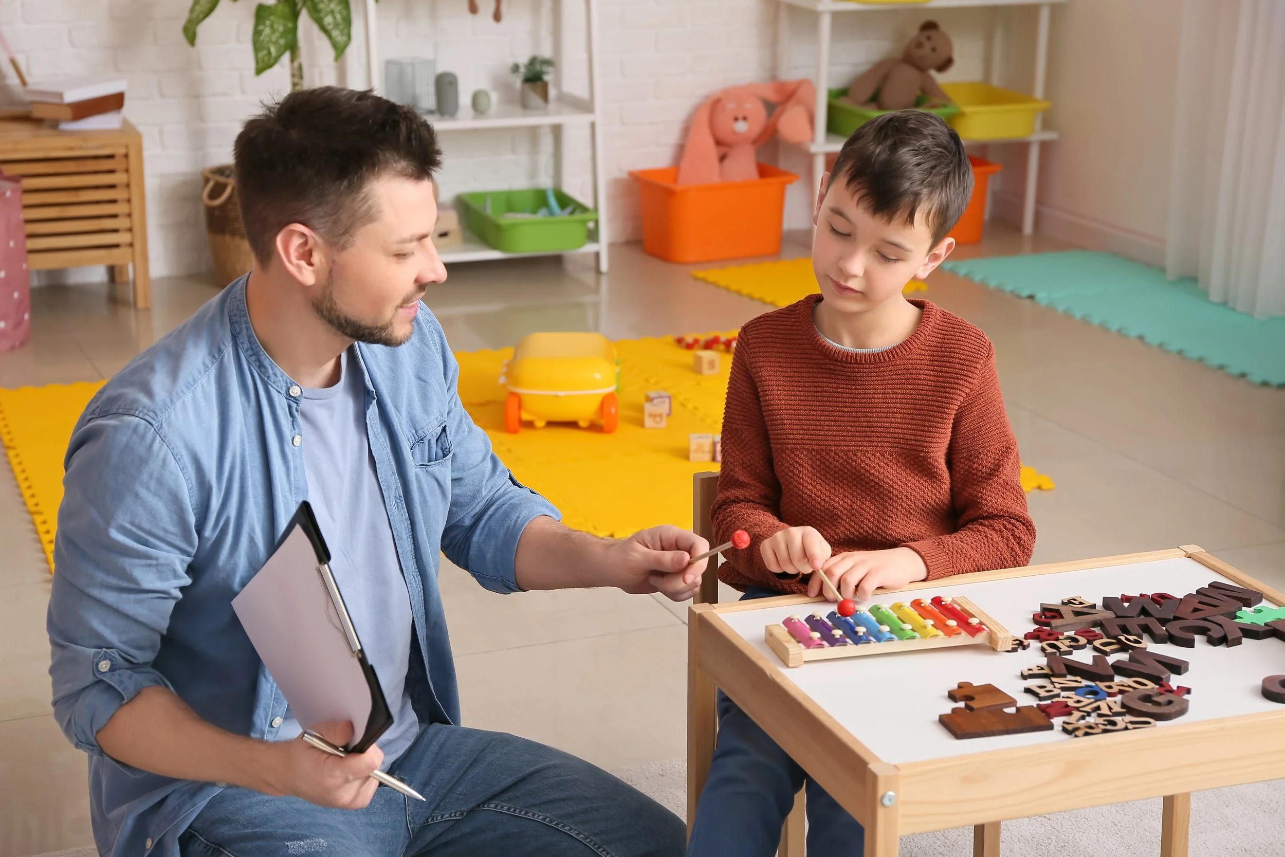 A boy and a man in a playroom with toys and colorful rugs, playing with a xylophone and a lollipop.