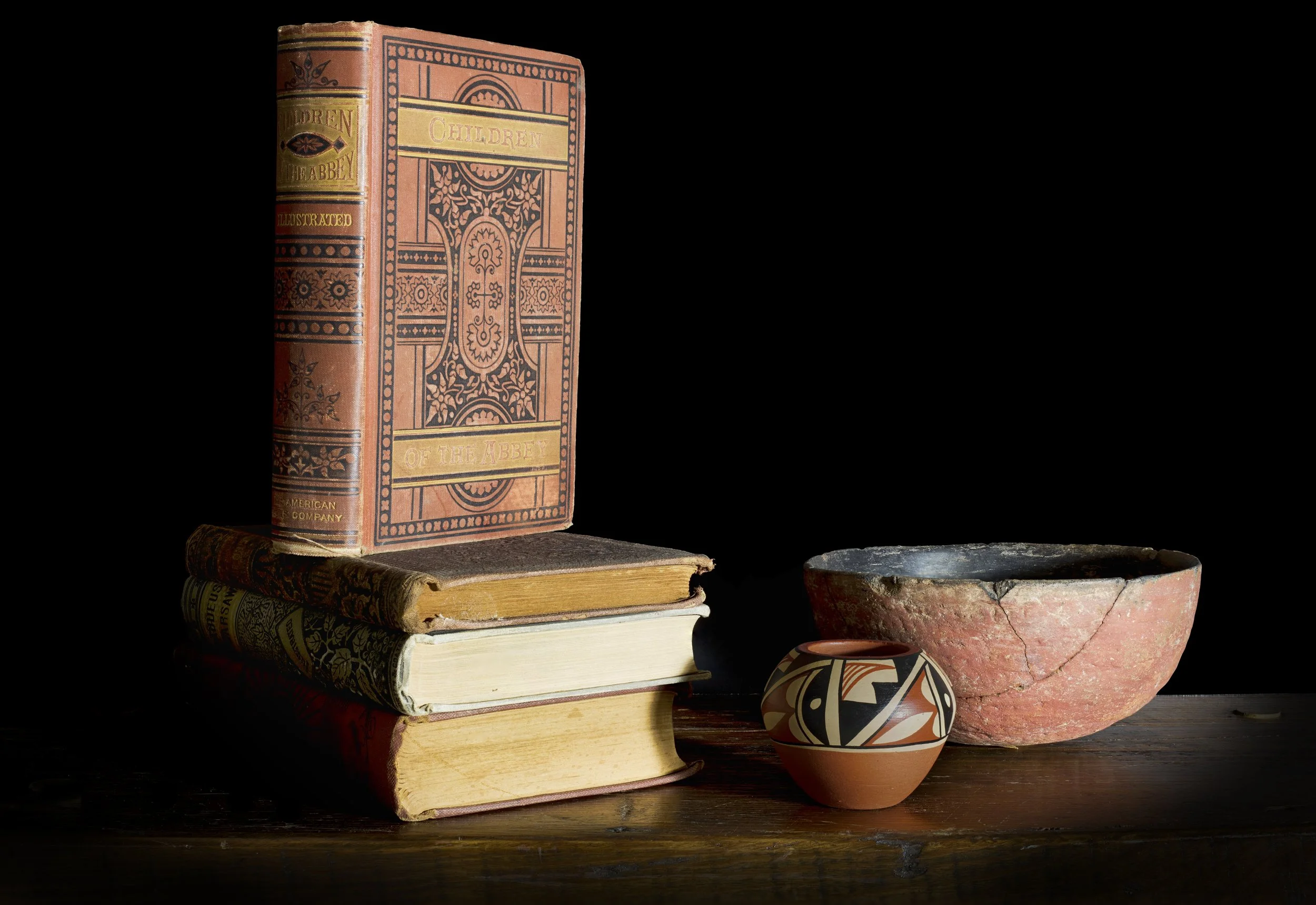 Stack of four books with decorative covers, a ceramic bowl, and a decorated pottery vessel on a wooden surface, dark background.