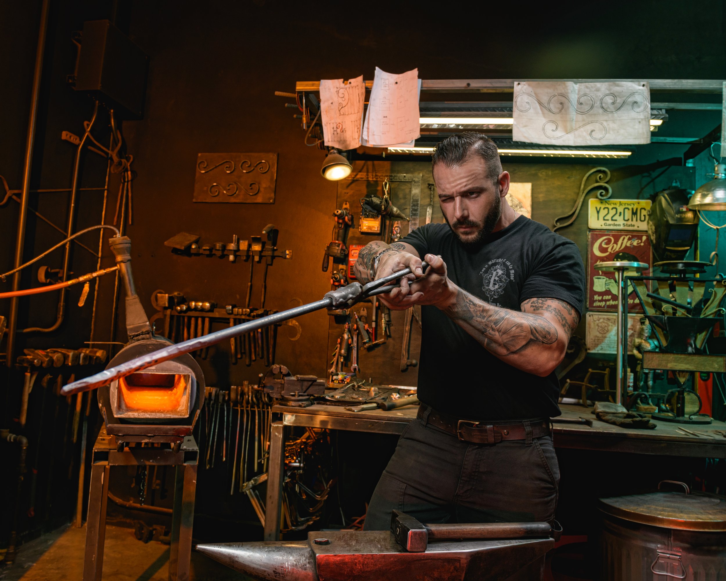 A man with tattoos working on a metal piece in a workshop filled with tools and equipment.