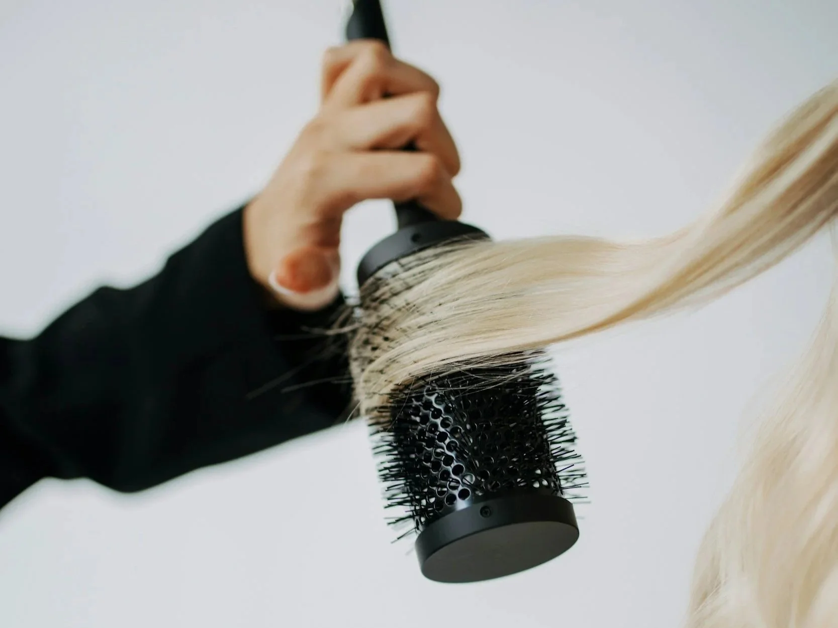 Close-up of a person styling blonde hair with a round brush and a curling iron.