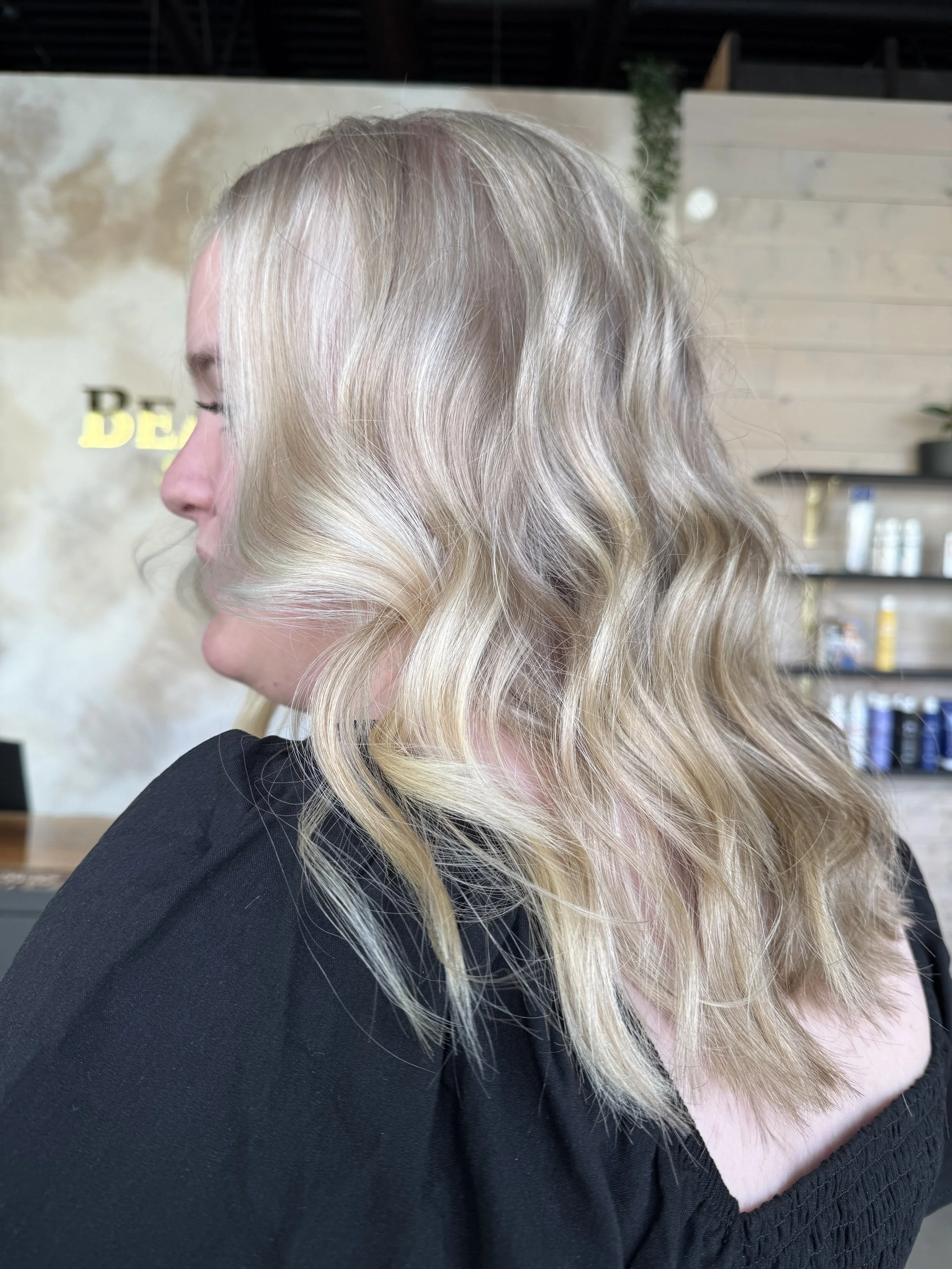 Close-up of a woman's side profile showing her long, wavy blonde hair with highlights, dressed in a black top, in a salon with blurred background.