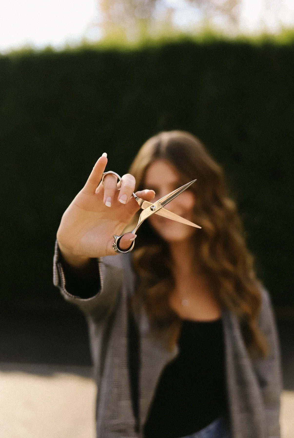 A woman holding a pair of gold scissors in front of her face, with the focus on the scissors and her face slightly blurred in the background.