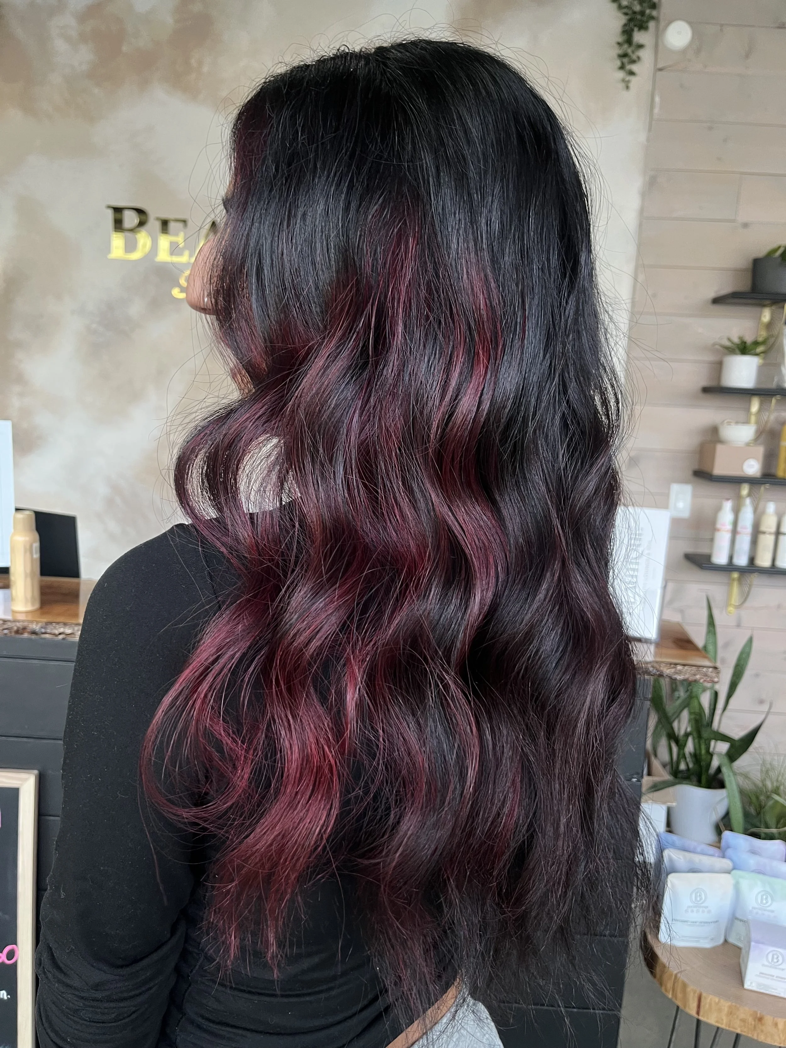 Side view of a woman with long, wavy black hair with pink highlights at the tips, standing in a hair salon.