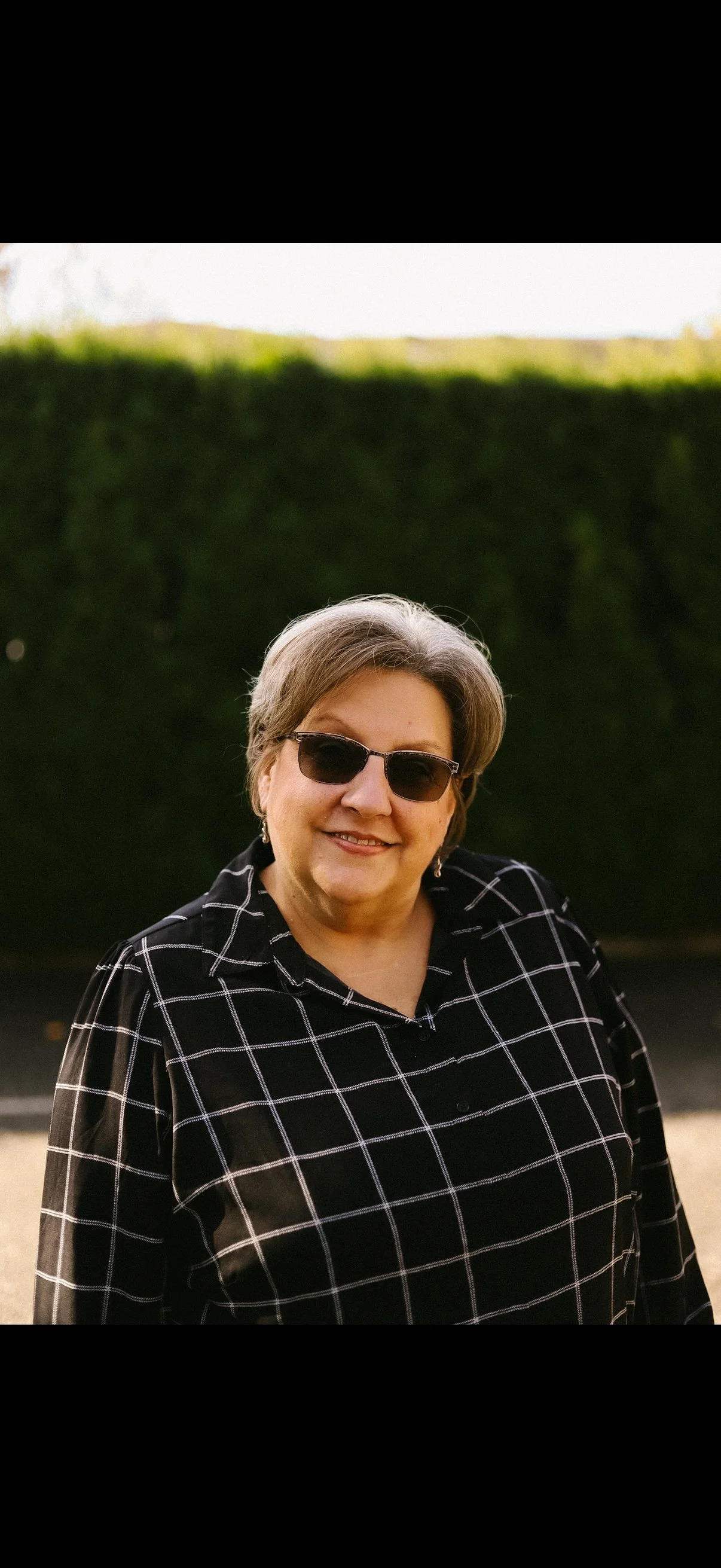 A smiling woman wearing dark sunglasses and a black and white checkered shirt outdoors.