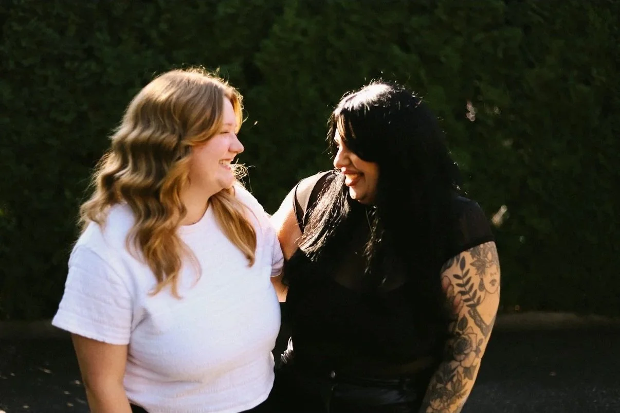 Two women smiling and looking at each other outdoors, one with blonde hair in waves wearing a white shirt, the other with black hair and tattoos wearing a black shirt, standing near a dark green hedge.