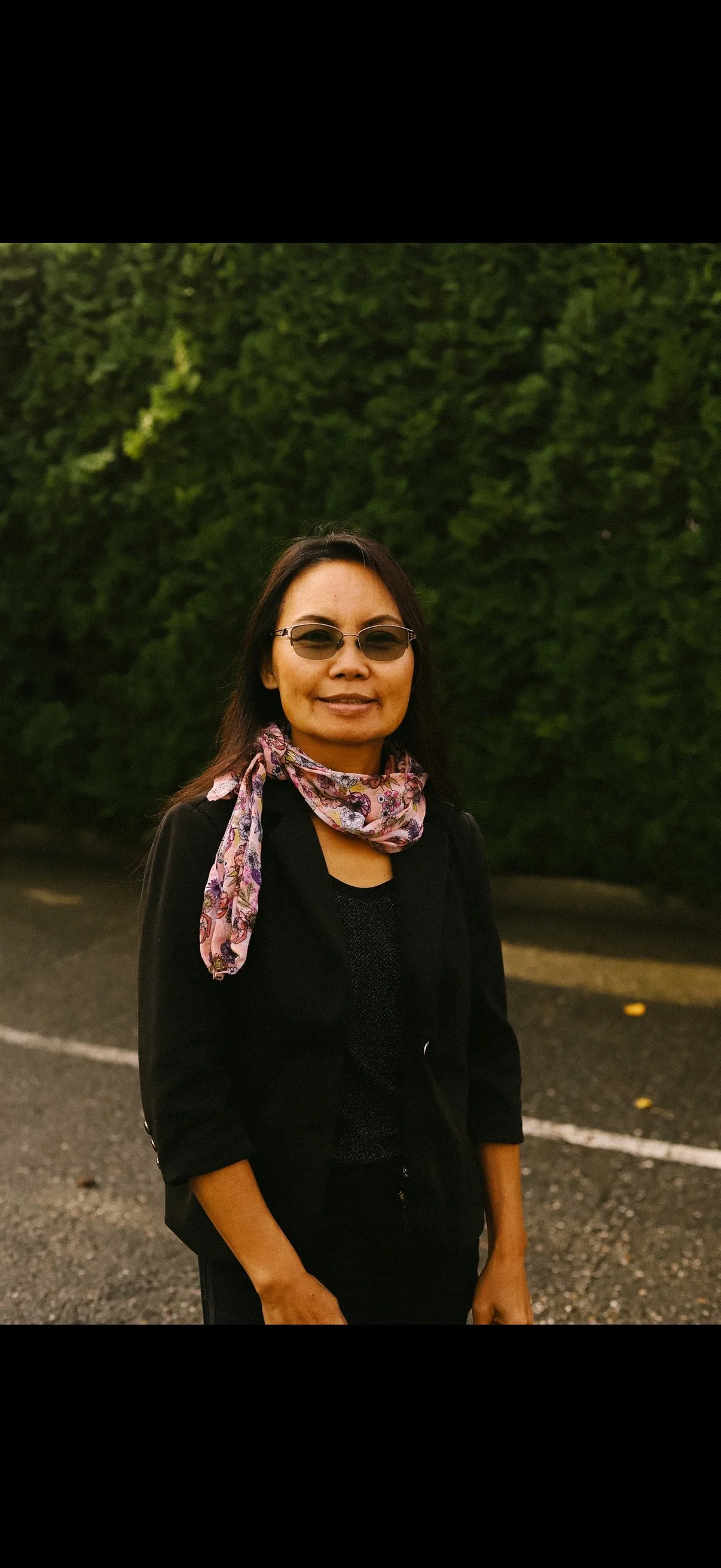A woman with black hair, wearing sunglasses and a floral scarf around her neck, standing outdoors in front of a green hedge.