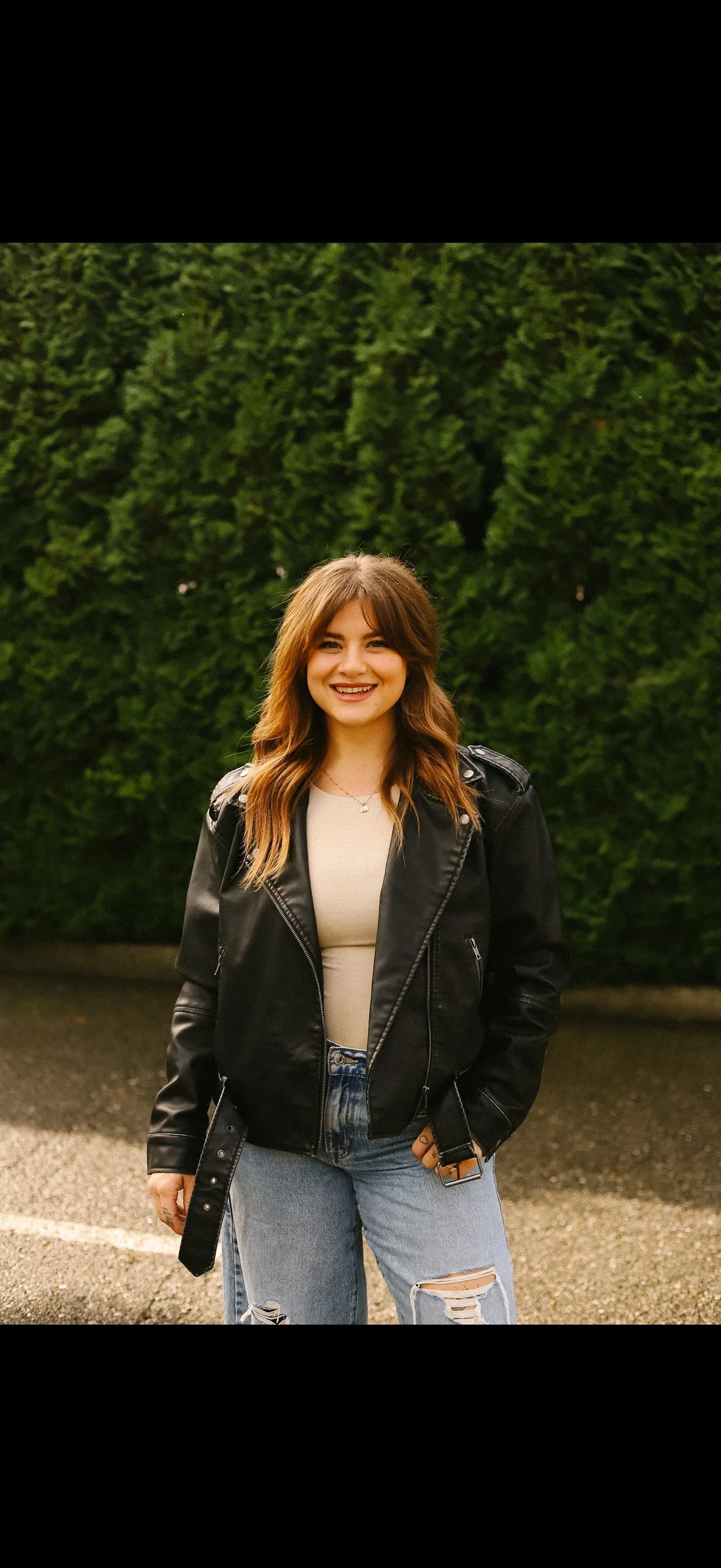 Young woman with brown hair smiling, wearing a black leather jacket, beige top, and ripped jeans, standing outdoors in front of green bushes.