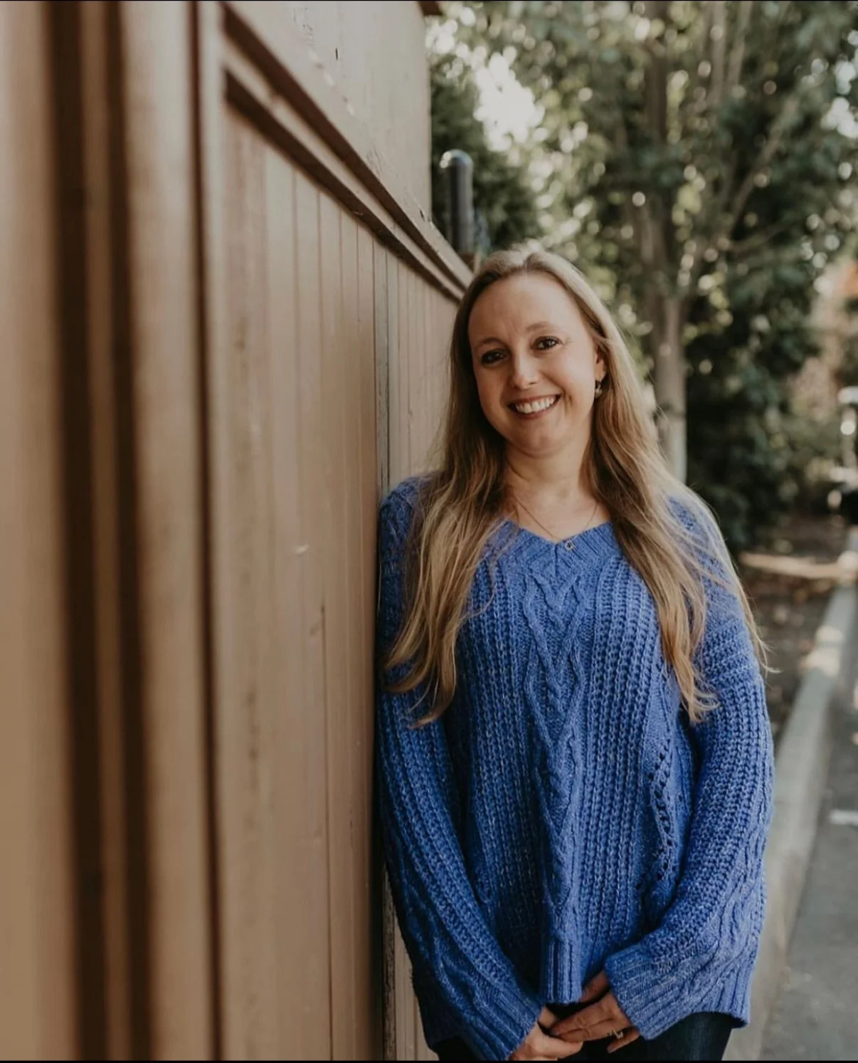 A woman with long blonde hair wearing a blue cable-knit sweater, smiling and leaning against a wooden fence outdoors.