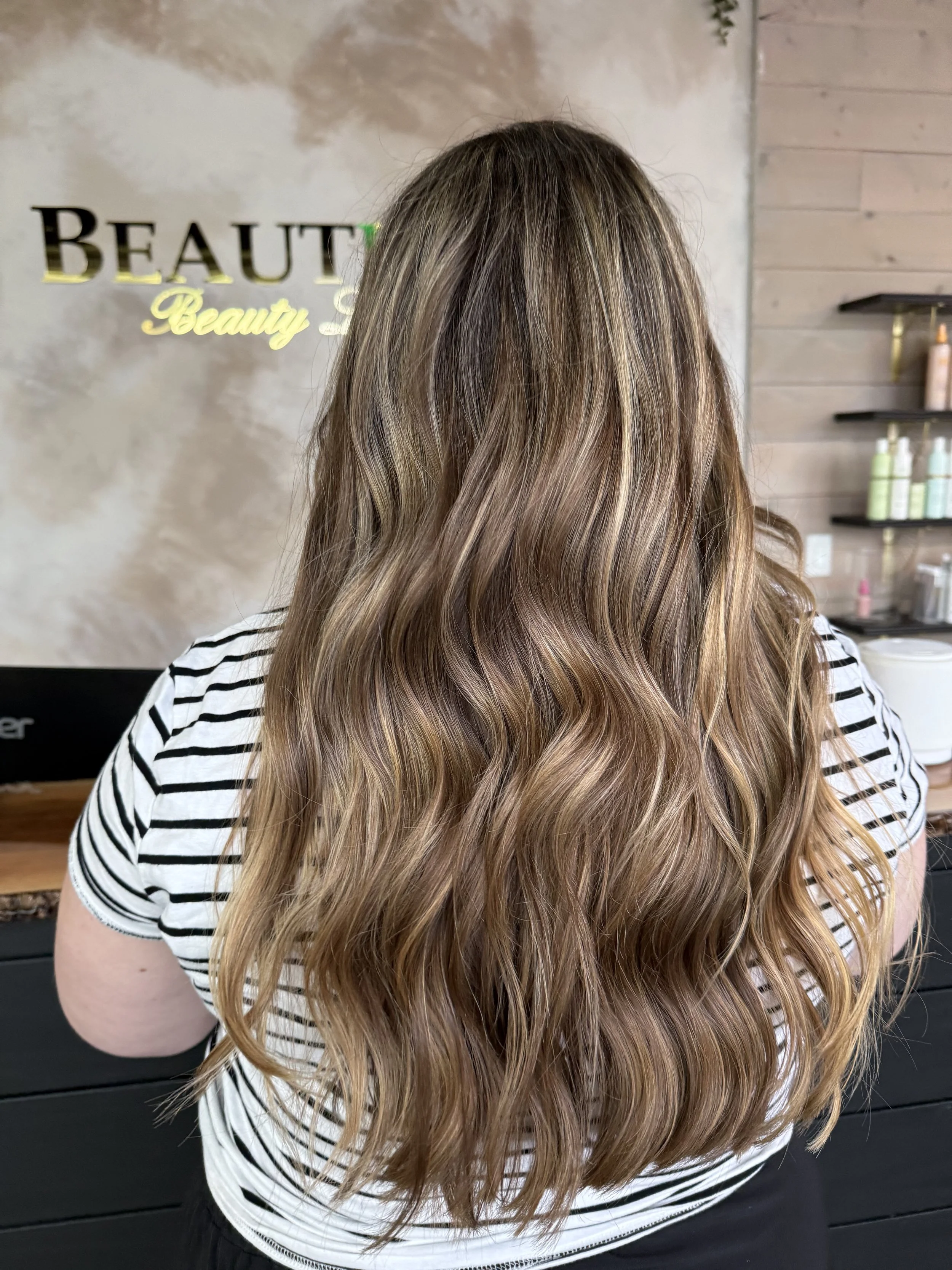 The image shows the back of a person with long, wavy, light brown hair with blonde highlights, standing in front of a wall with the words 'BEAUTY' and 'Beauty Spa'. The person is wearing a black and white striped shirt.
