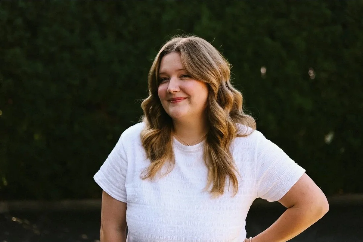A woman with light brown, wavy hair is standing outdoors in front of dark green foliage, wearing a white t-shirt and smiling slightly.
