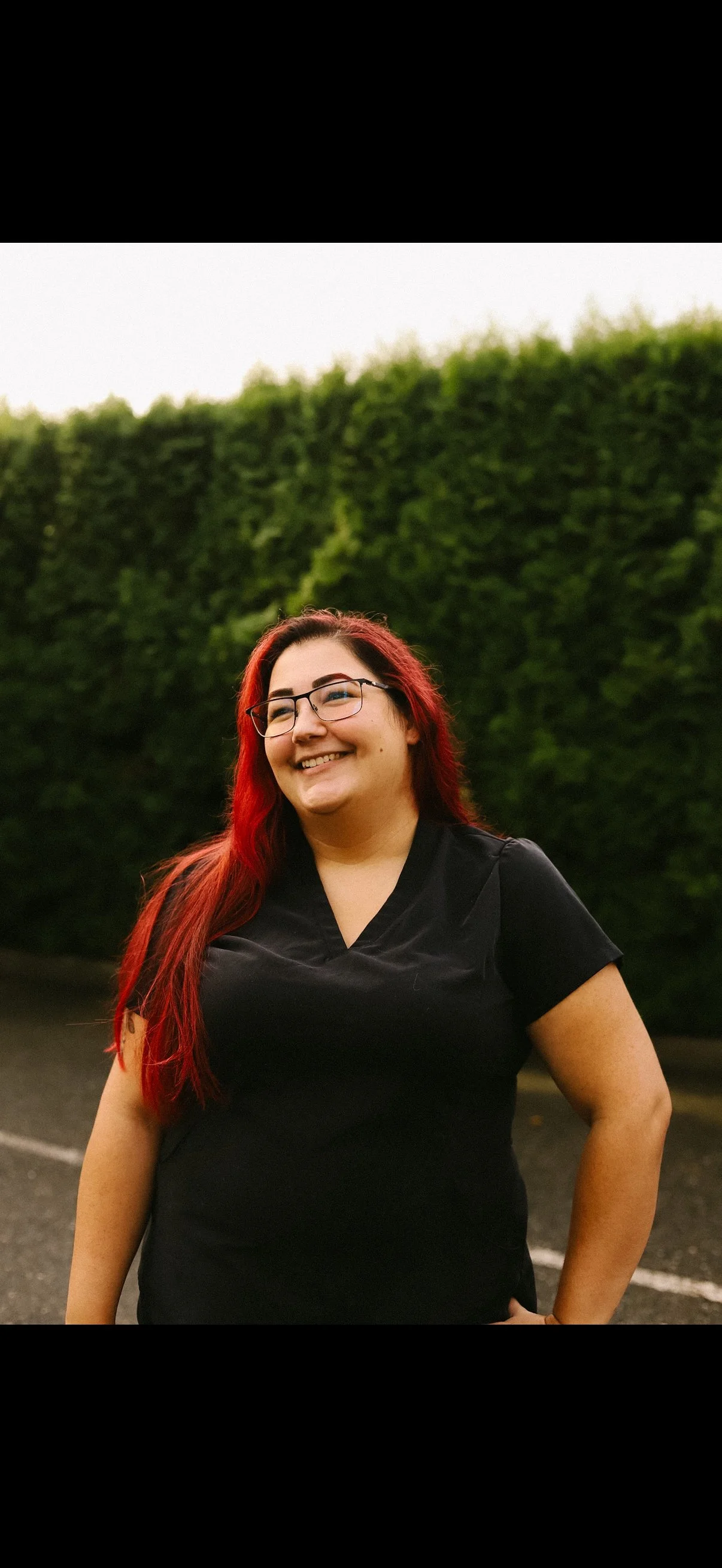 A woman with long red hair, glasses, and a black shirt smiling outdoors with green trees in the background.