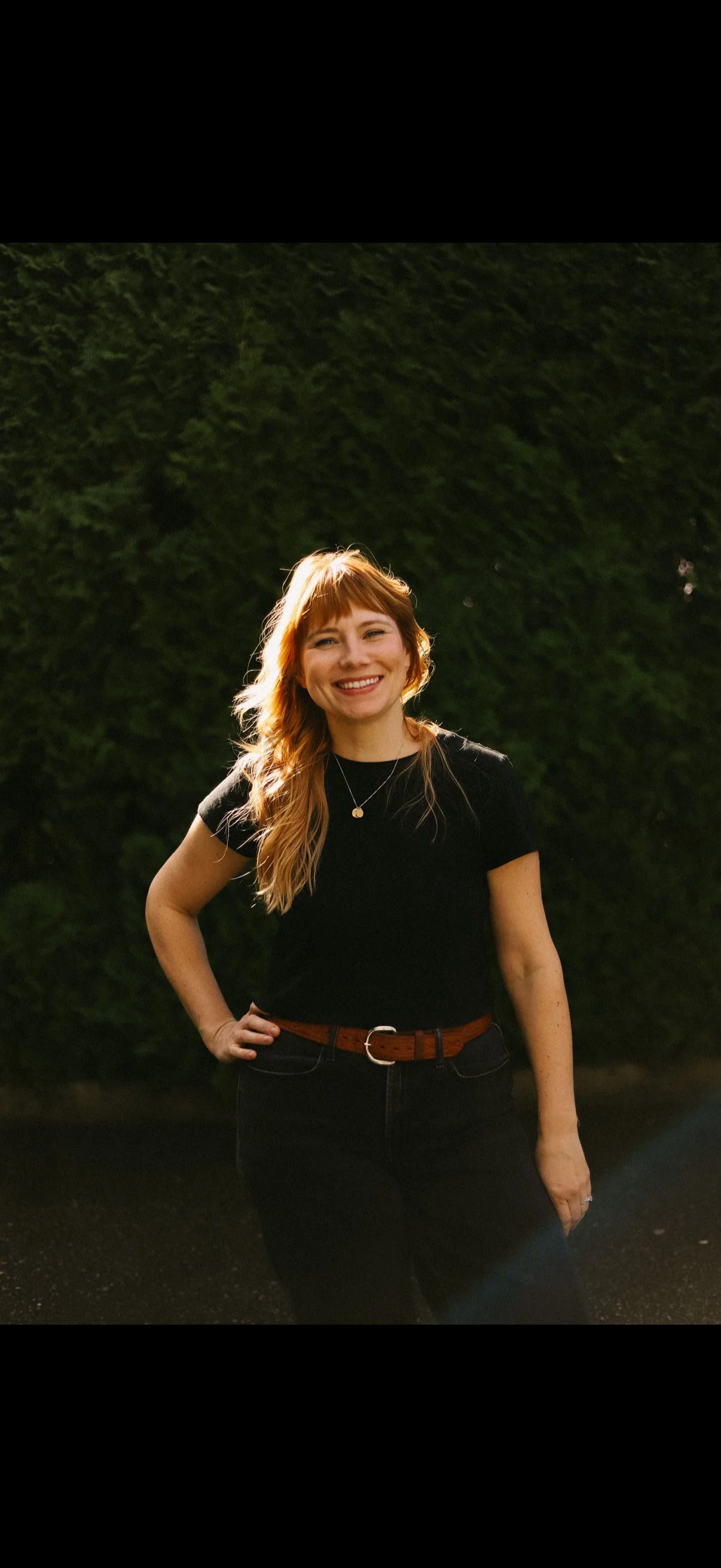 A young woman with ginger hair smiling, wearing a black t-shirt, black jeans, and a brown belt, standing outdoors in front of a green hedge.