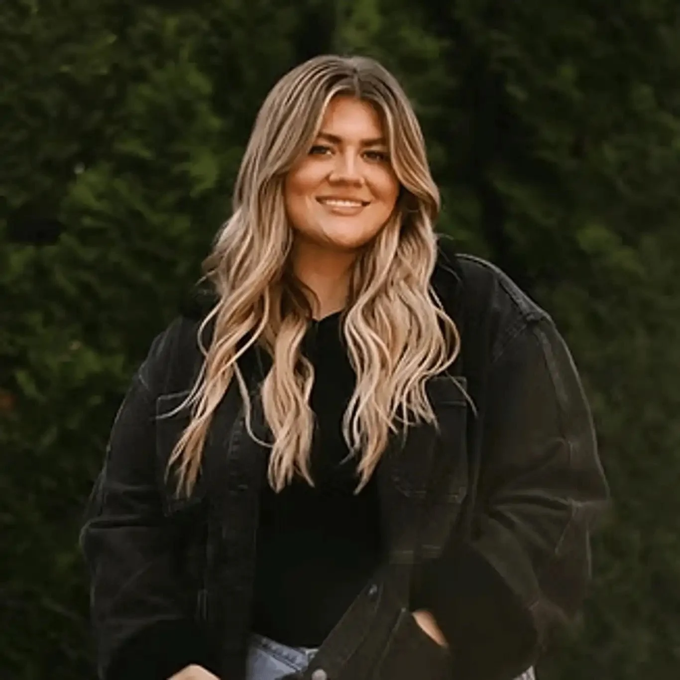 A young woman with long wavy blonde hair smiling outdoors with green trees in the background, wearing a black jacket.