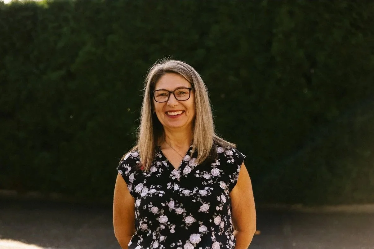 A woman wearing glasses and a black floral blouse standing outdoors in front of a dark green hedge, smiling at the camera.