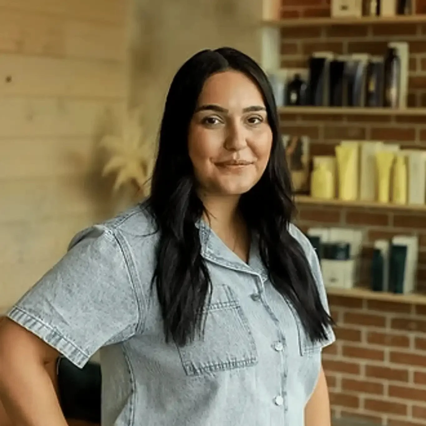 A woman with long black hair wearing a light denim shirt, standing in a room with brick walls and a bookshelf.