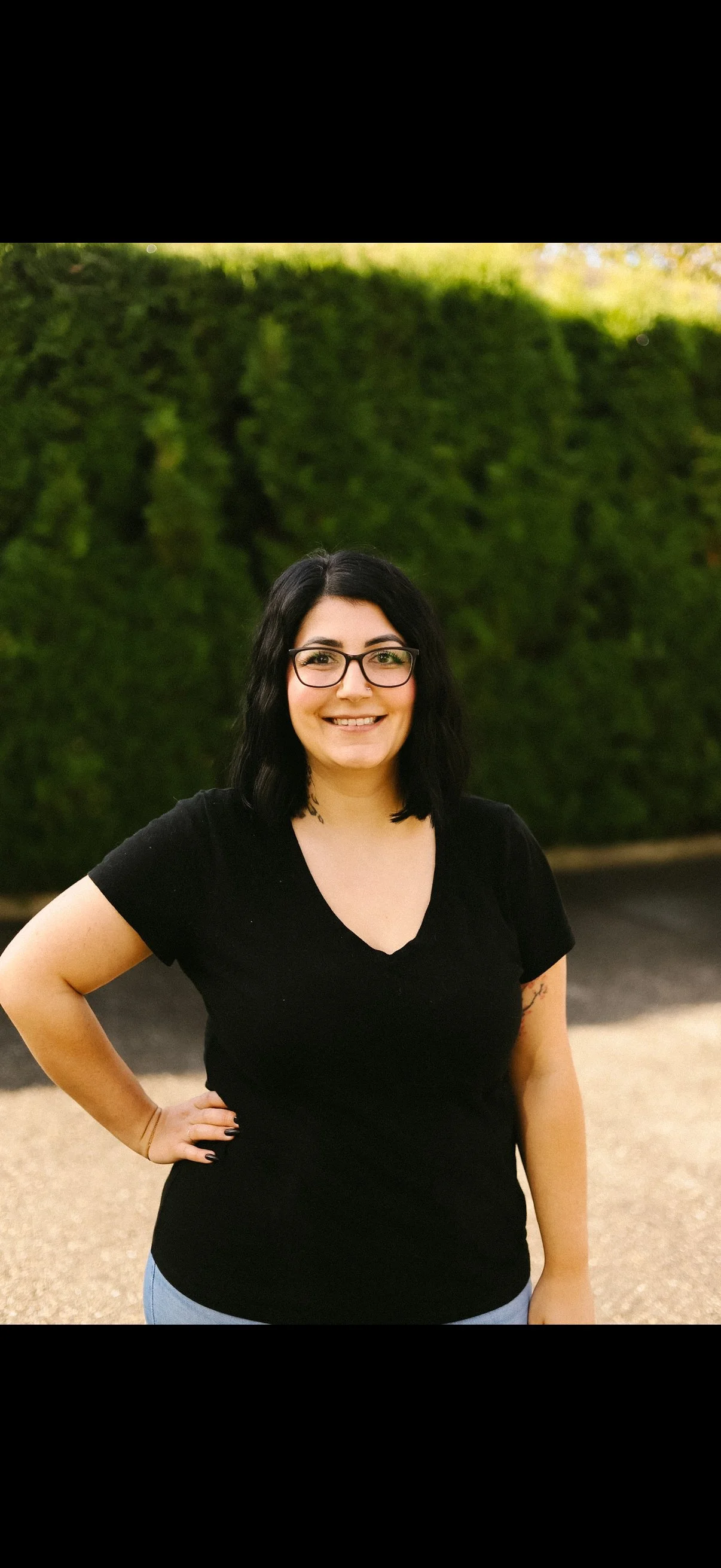 A woman with shoulder-length black hair, glasses, and a black T-shirt smiling outdoors with a green hedge in the background.