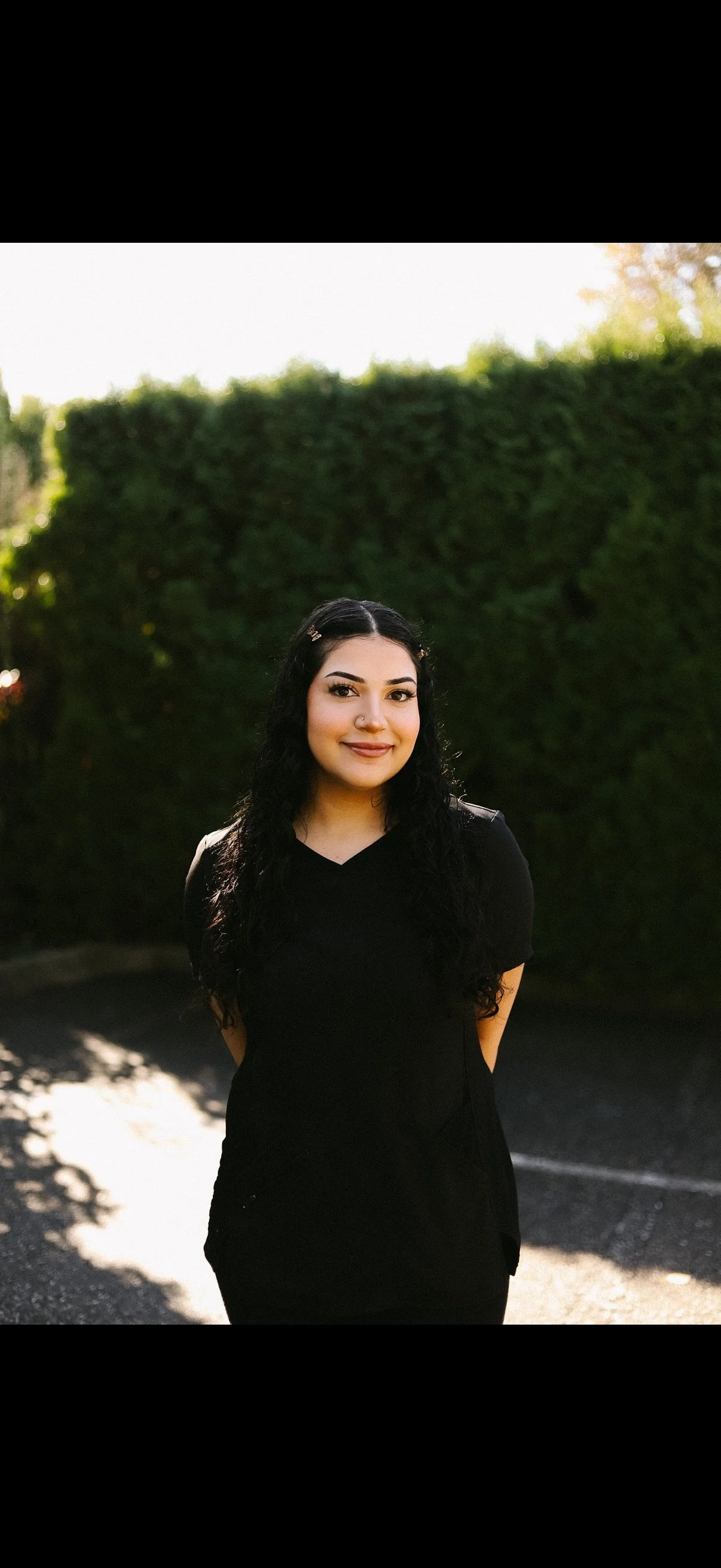 A young woman with long, curly black hair, wearing a black top, standing outdoors in front of a green hedge, smiling at the camera with sunlight casting shadows on the ground.