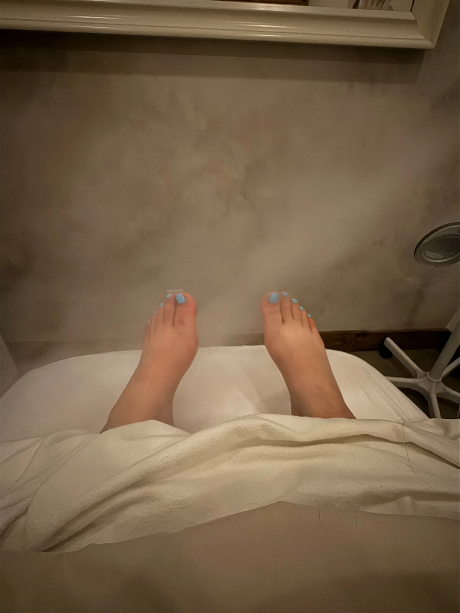 A person with blue painted toenails standing on a steam room floor, viewed from above, with part of their white hospital gown visible.