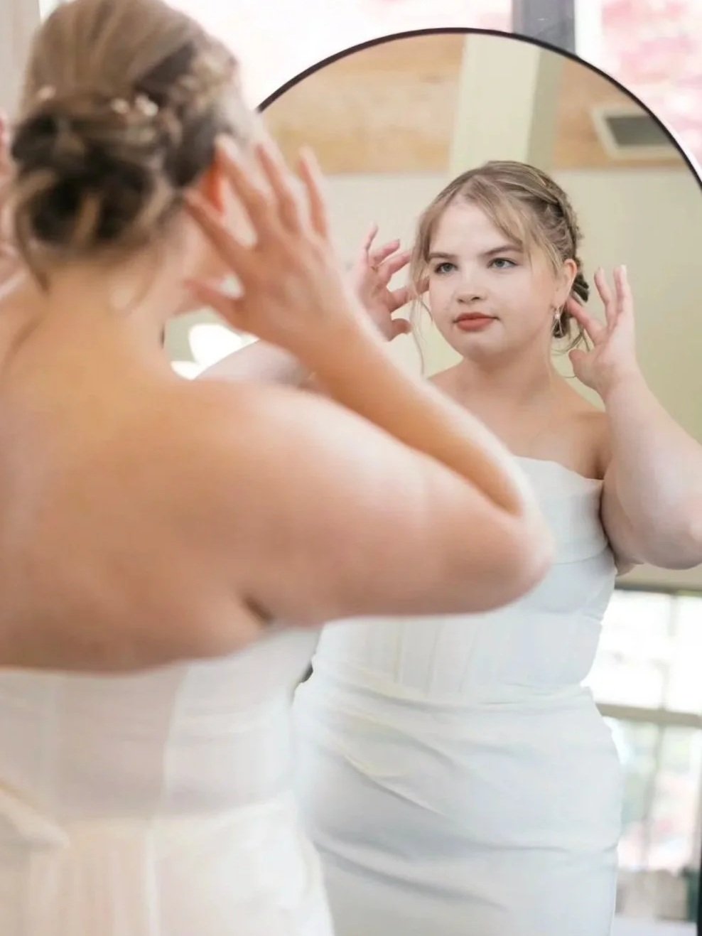 A woman with styled hair is looking at her reflection in a round mirror. She is adjusting her hair and wearing a white strapless dress or top, with a soft indoor background.