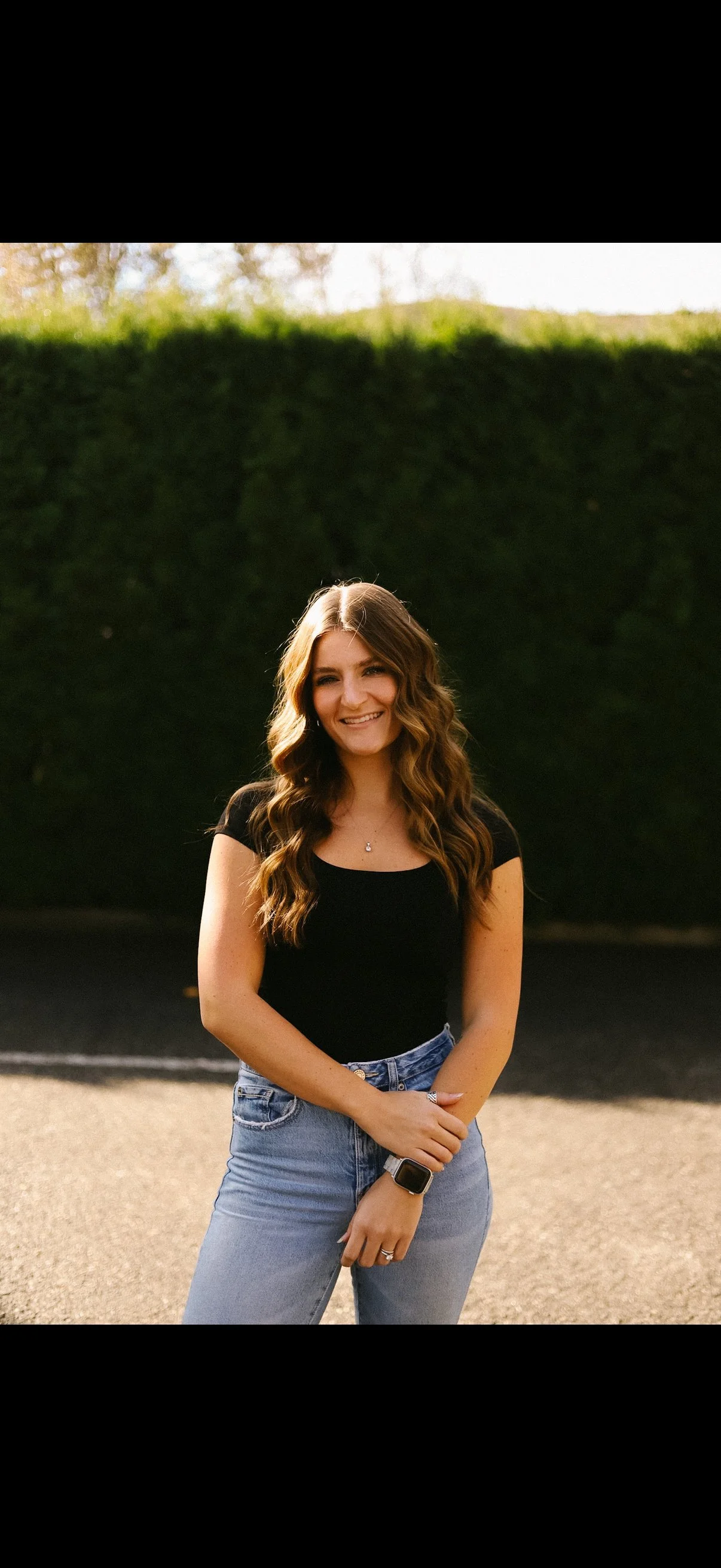 A young woman with long, wavy brown hair, wearing a black t-shirt, light blue jeans, a watch, and a ring, smiling outdoors with a dark hedge and sunny sky in the background.