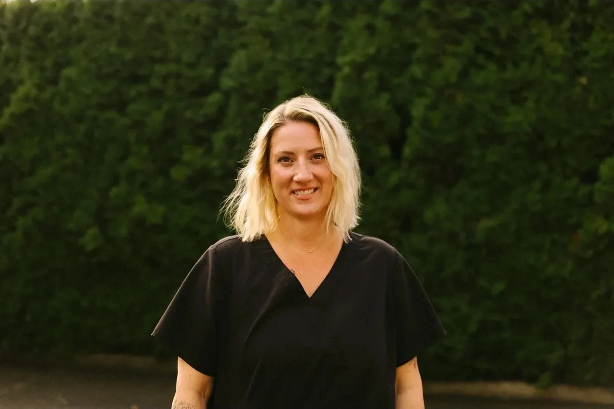 A woman with blonde hair wearing a black shirt standing outdoors in front of green foliage.