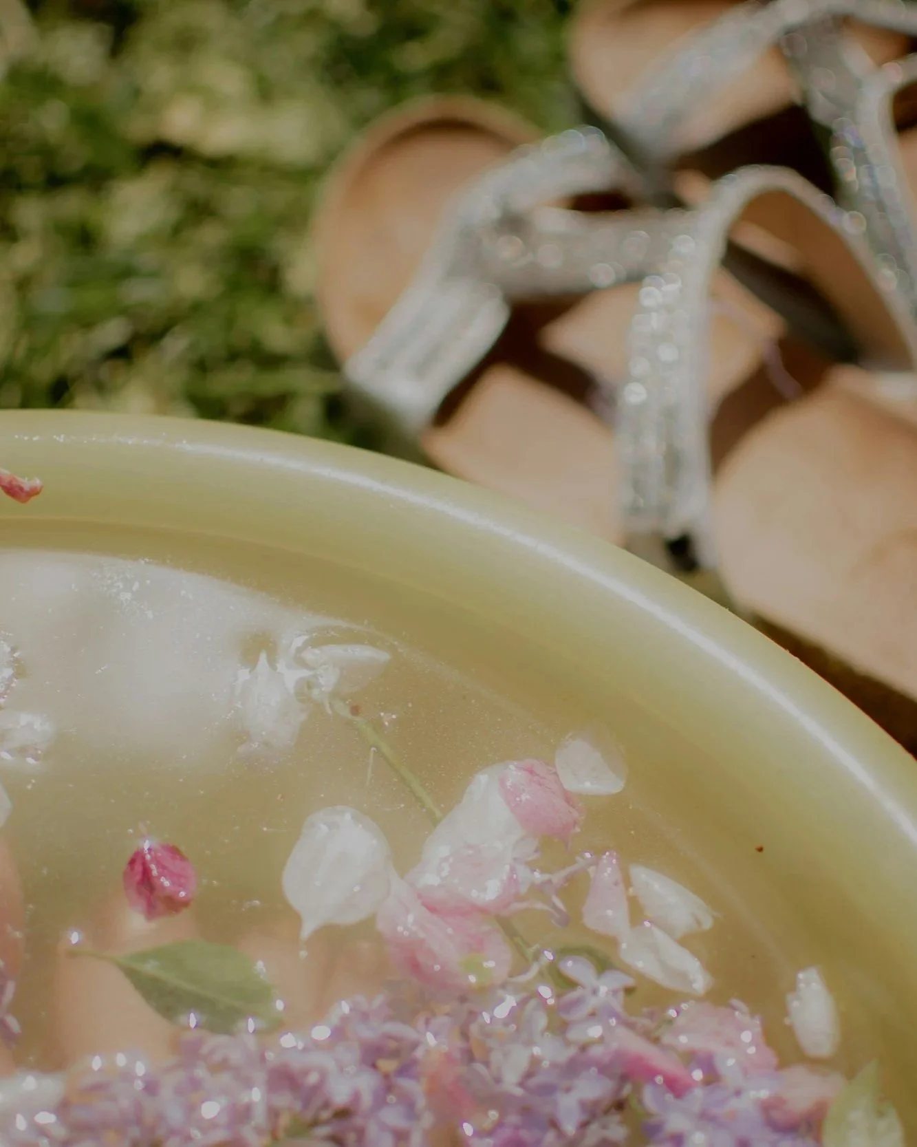 A yellow candle with flower petals and small leaves floating on melted wax near a pair of silver, glittery high-heeled sandals with ankle straps on grass.