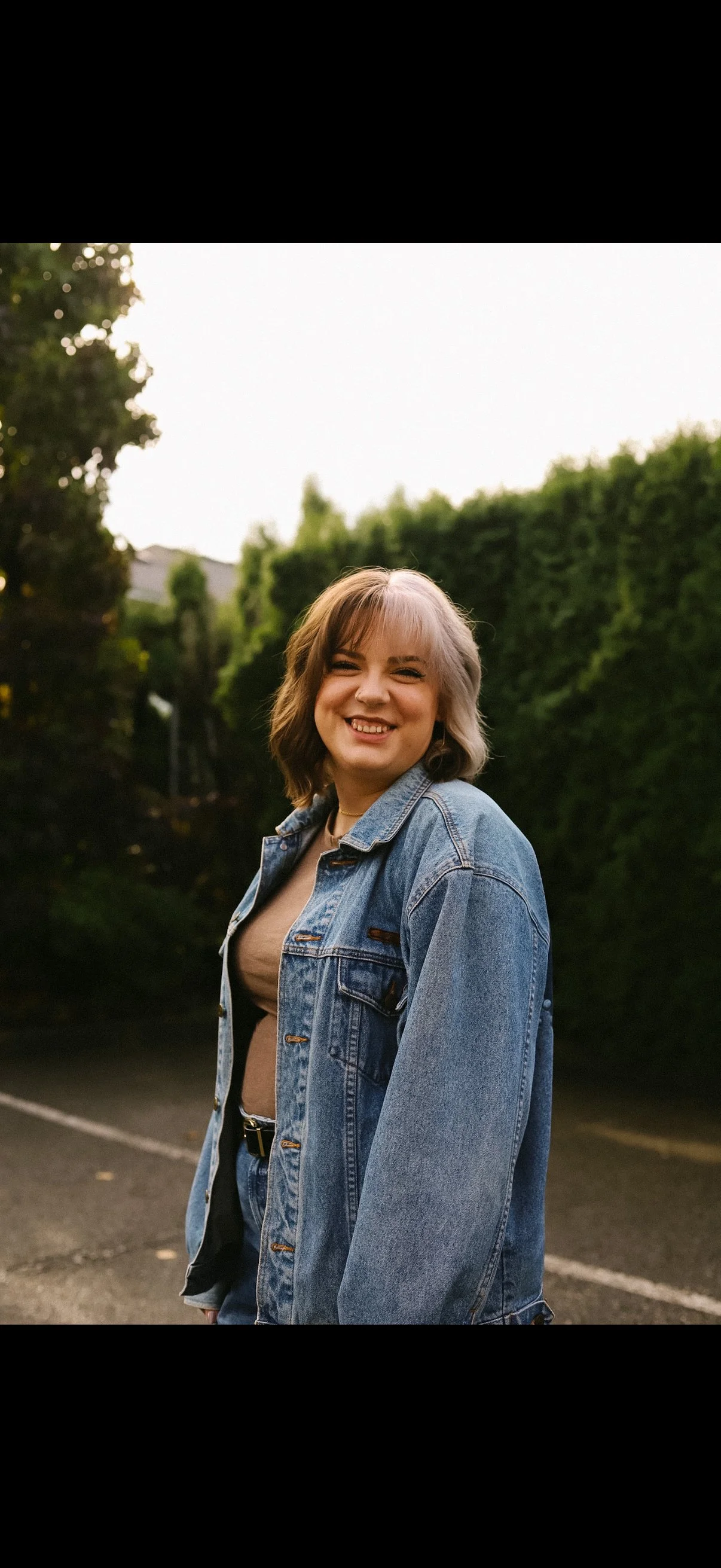A young woman smiling outdoors in a parking lot, wearing a denim jacket over a beige top, with greenery in the background during the evening.