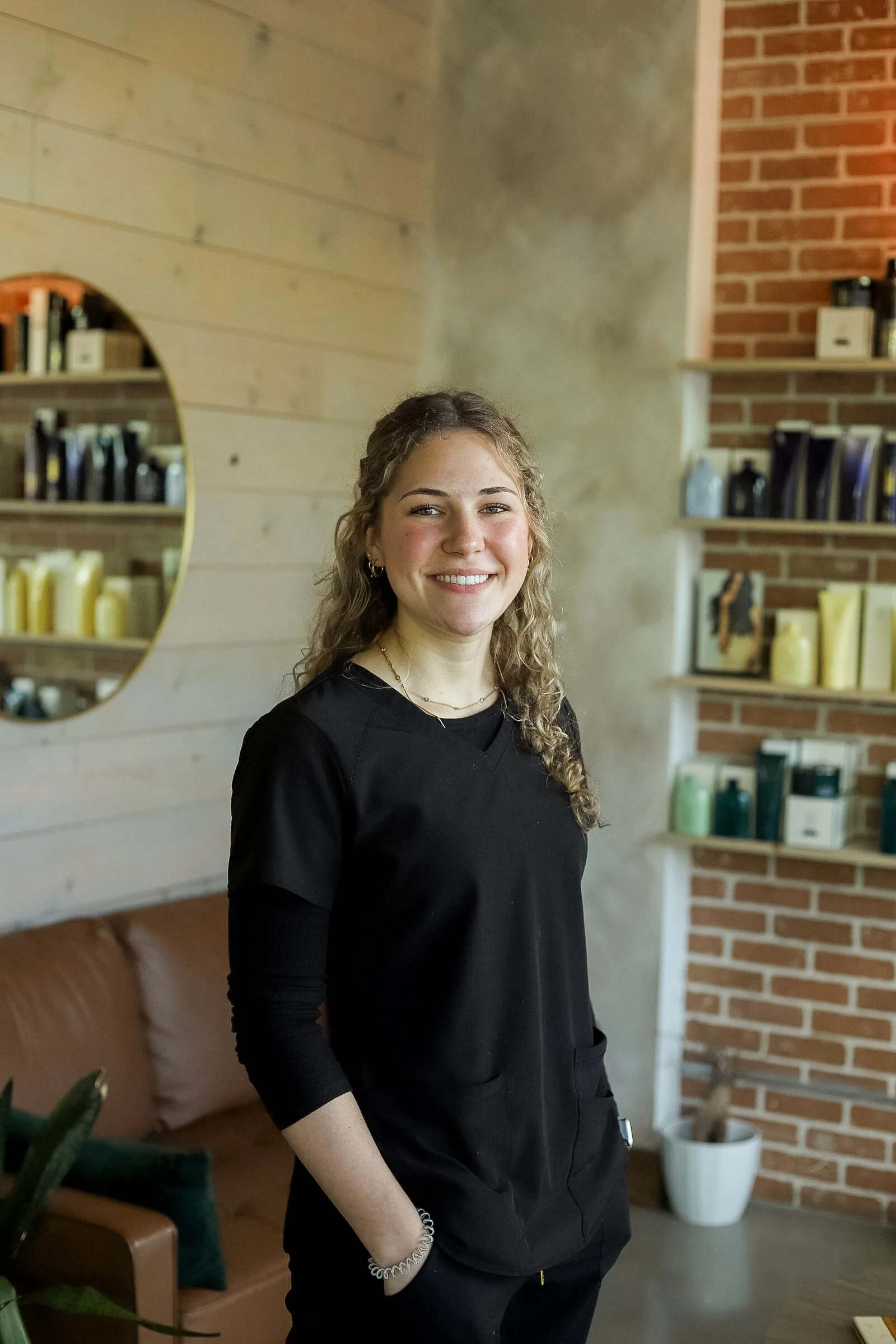 A young woman with curly blonde hair smiling and standing in a modern interior space with a wooden wall, a round mirror, a brick wall shelf with products, and a brown leather couch.