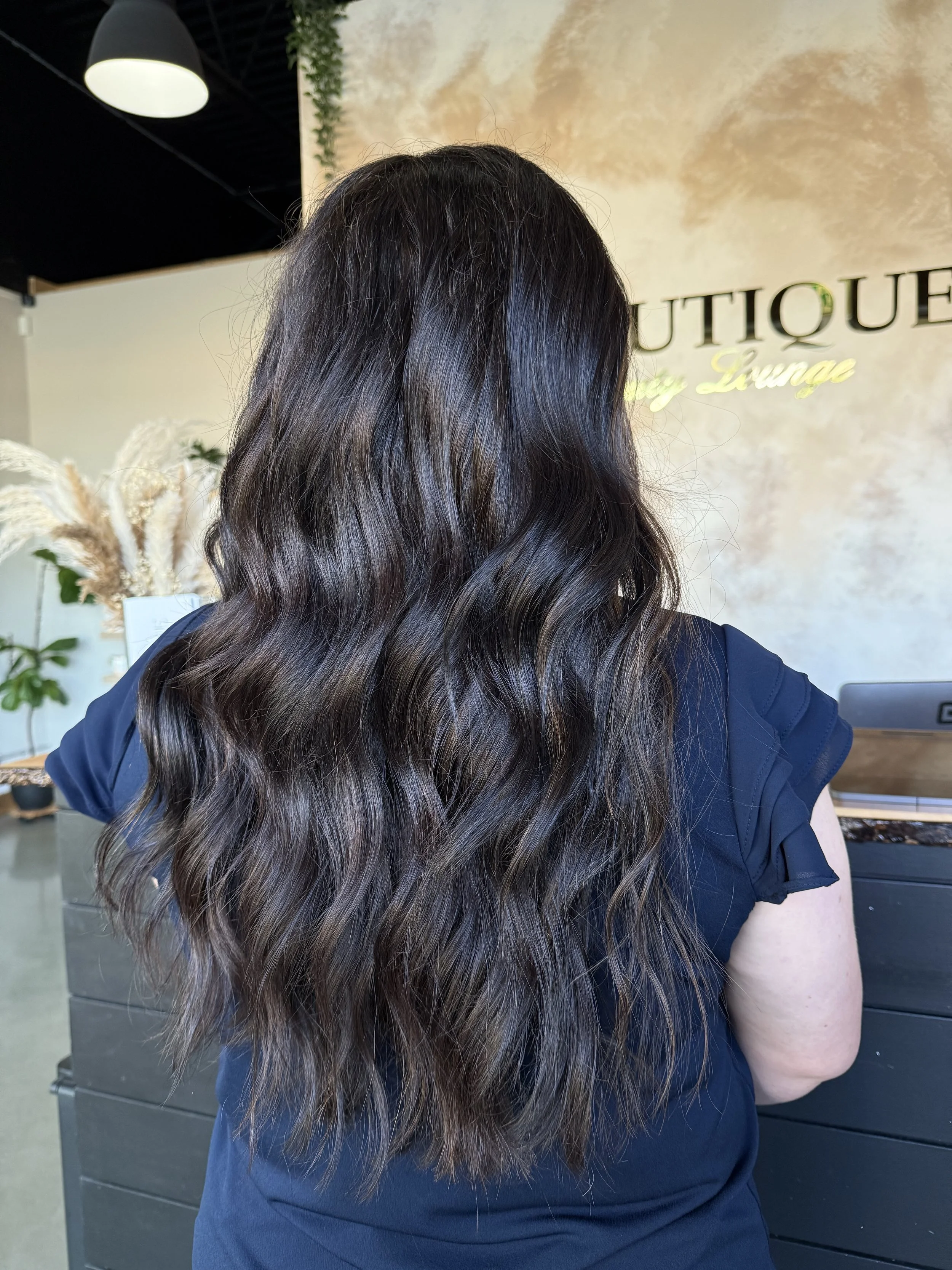A woman with long wavy dark hair with lighter highlights standing in front of a beige wall with the words 'Boutique Lounge' on it, taking a photo or looking at a screen.