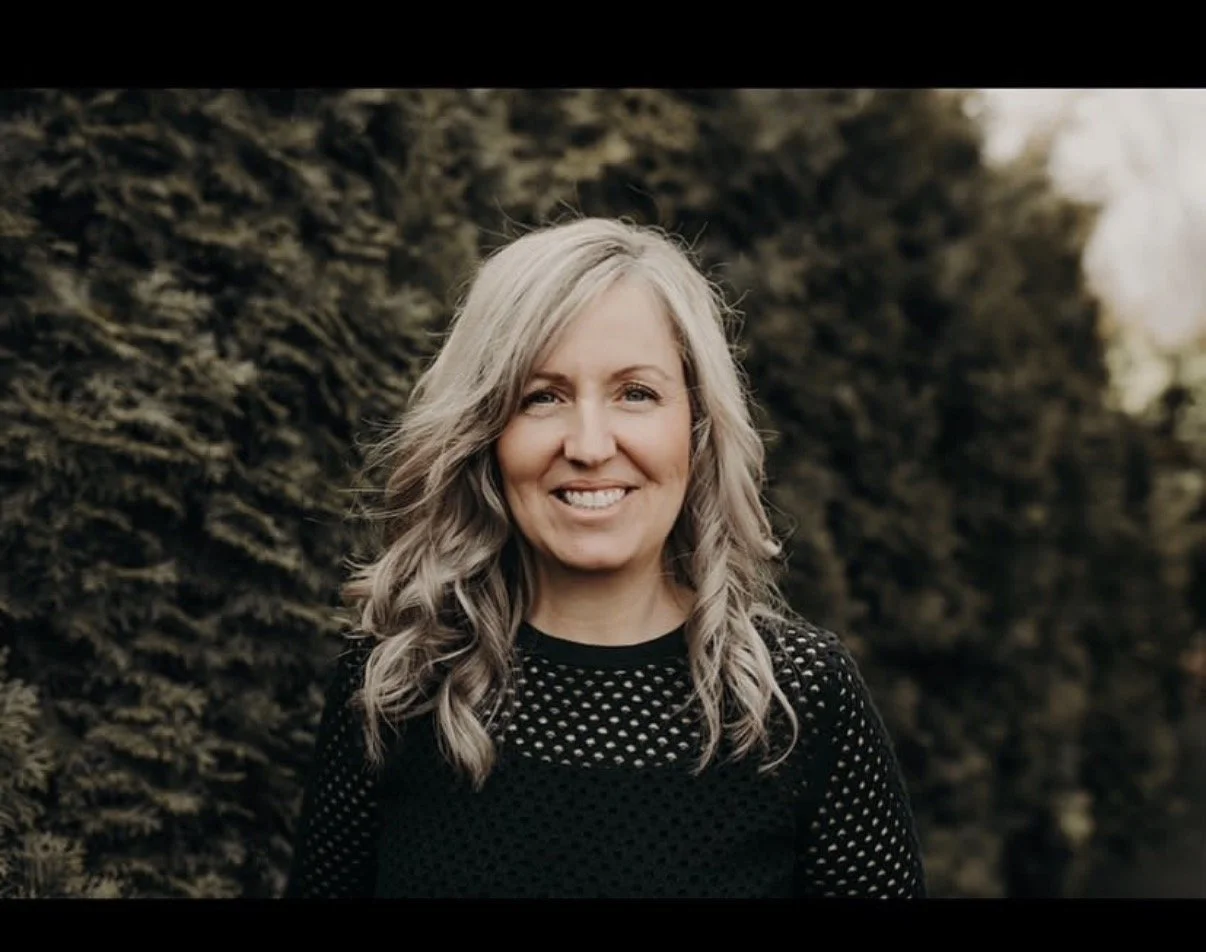 A woman with silver hair smiling outdoors, standing in front of a hedge.