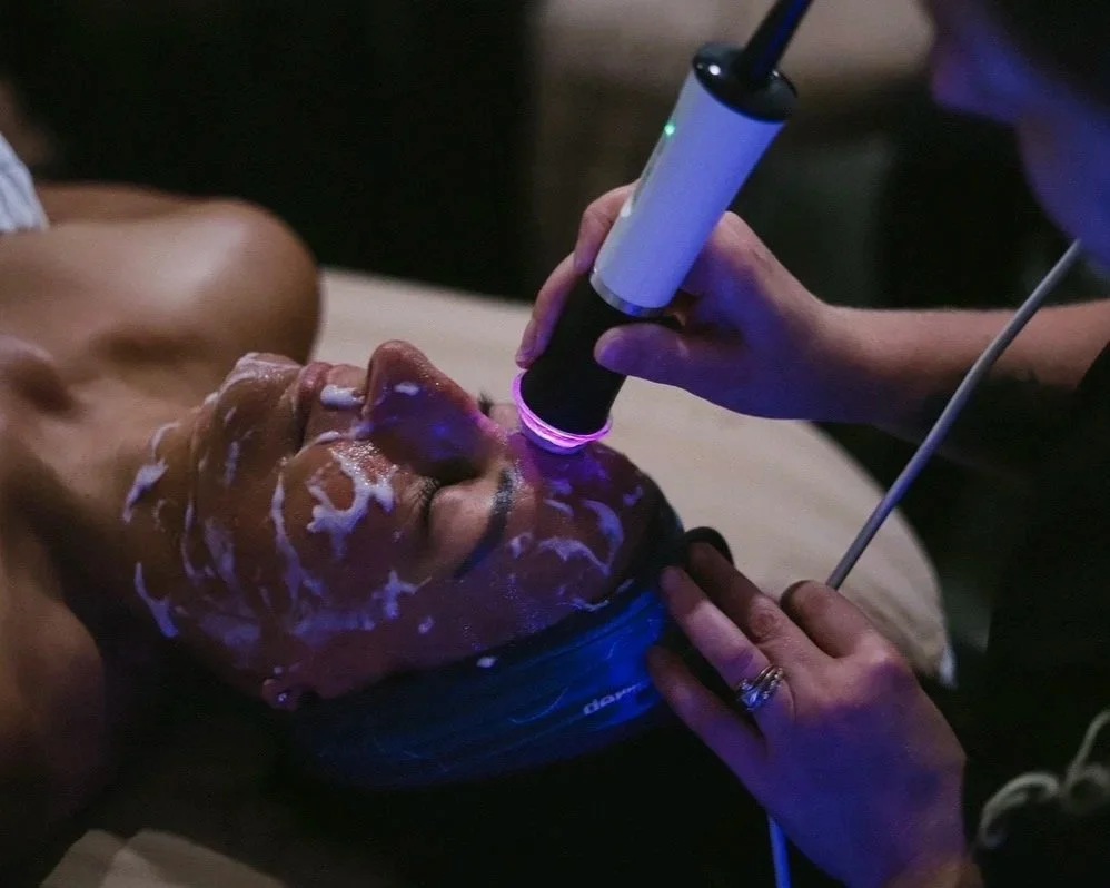 A person receives a facial treatment with a handheld laser device from a professional in a dark room.