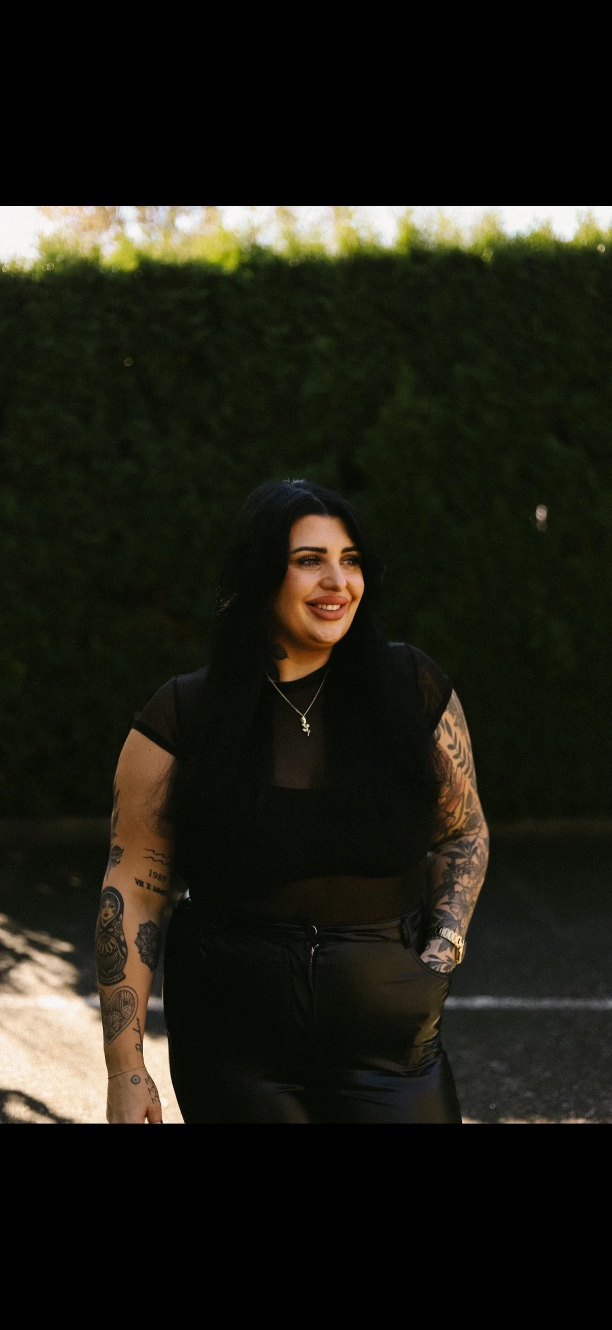 A woman with long black hair and tattoos on her arms, standing outdoors near a beach with a hedge in the background, smiling and wearing black clothing.