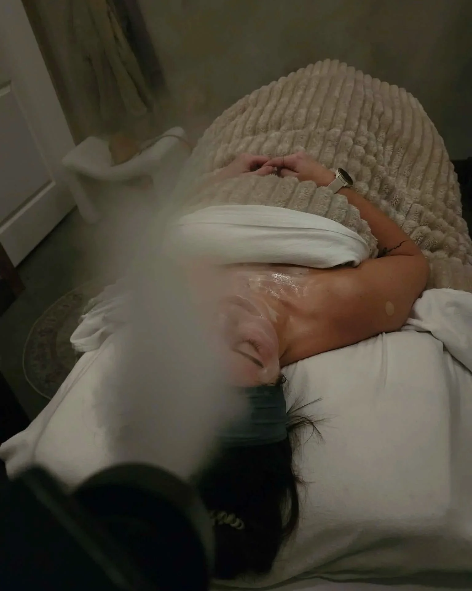 A woman lying on a bed covered with a beige blanket, resting with her eyes closed, and wearing a white shirt and watch, in a dimly lit room.