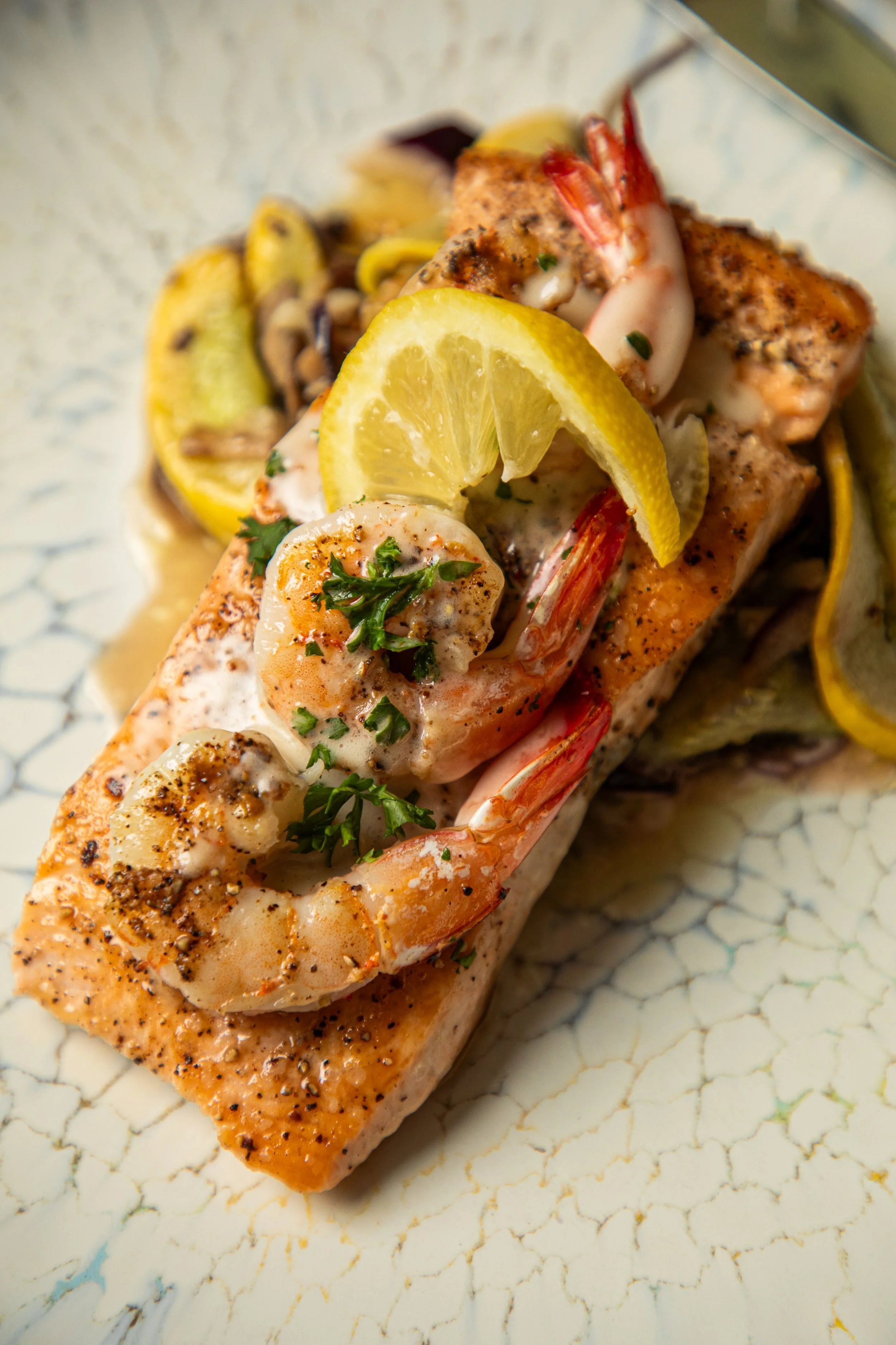 Close-up of a grilled salmon topped with lemon slices, shrimp, herbs, and seasoning, on a patterned white plate.