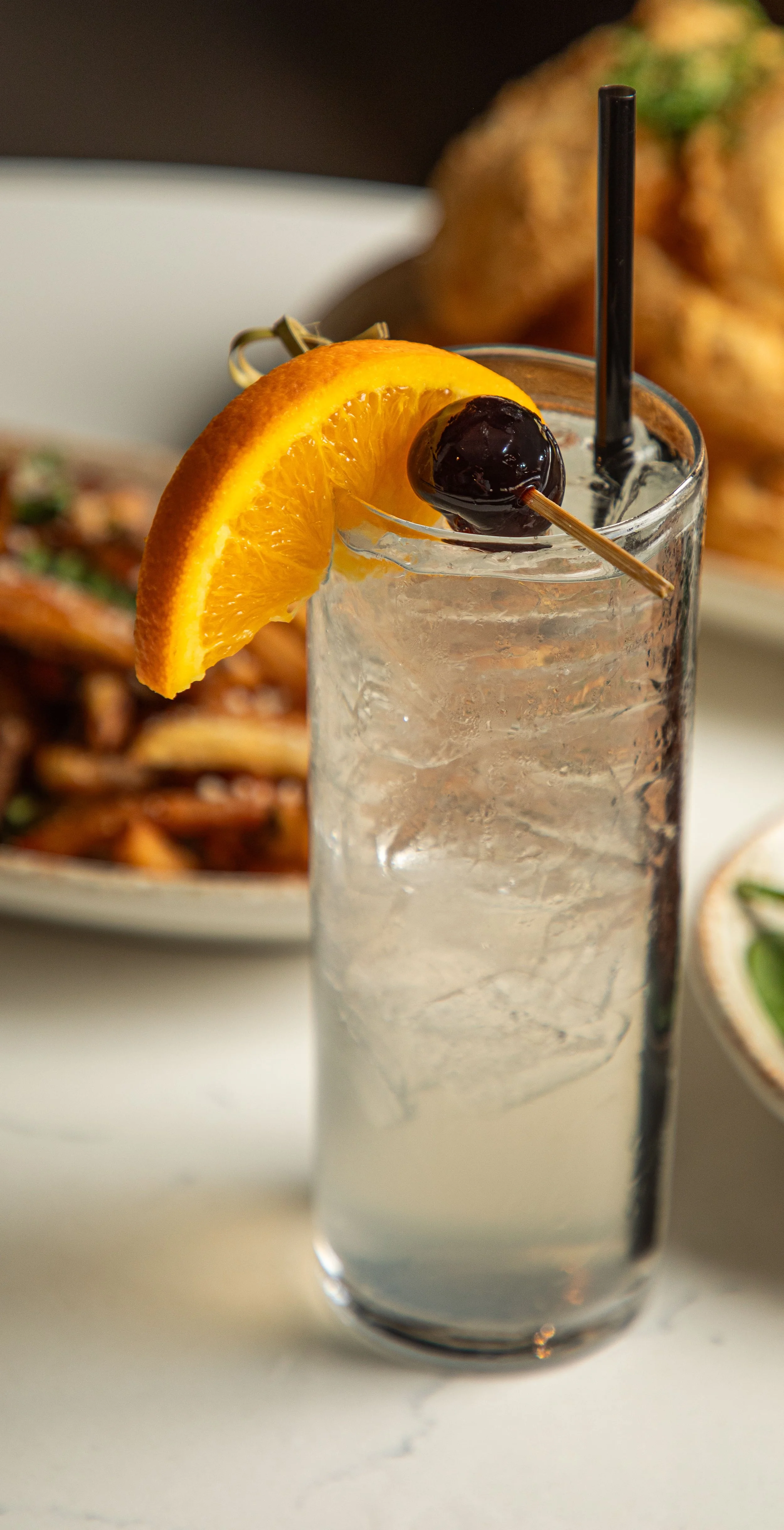A tall glass of clear soda with ice, garnished with an orange slice and a black cherry on a pick. There are plates of food in the background.