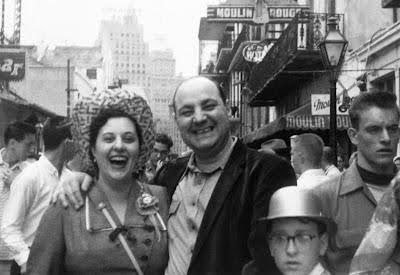 A black and white photo of several people on a street, including a woman with curly hair and a man smiling, with buildings and street signs in the background.