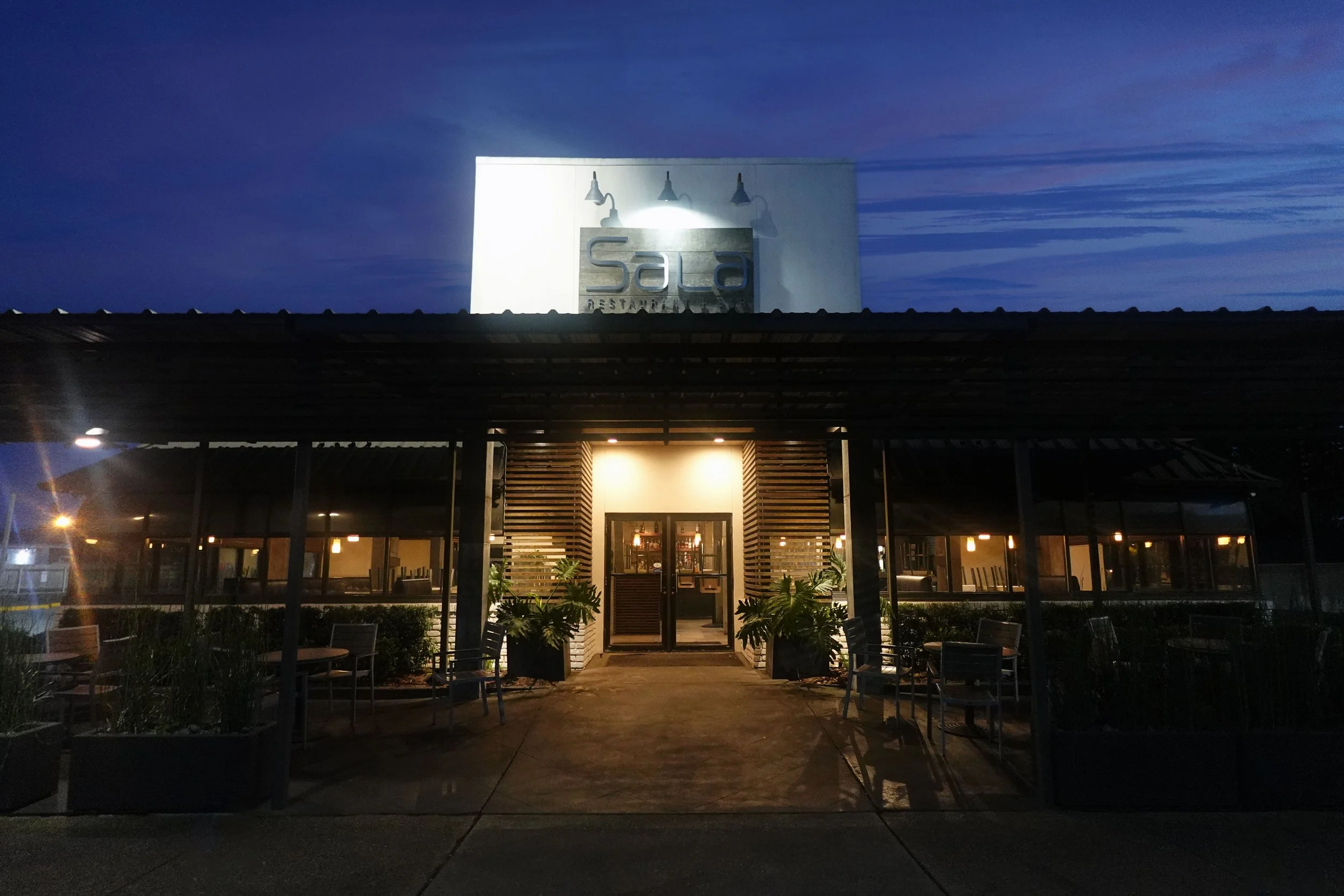 Nighttime view of the exterior of a restaurant called Sala, with illuminated sign on a white building, outdoor seating area with chairs and tables, and some plants, under a dark sky.