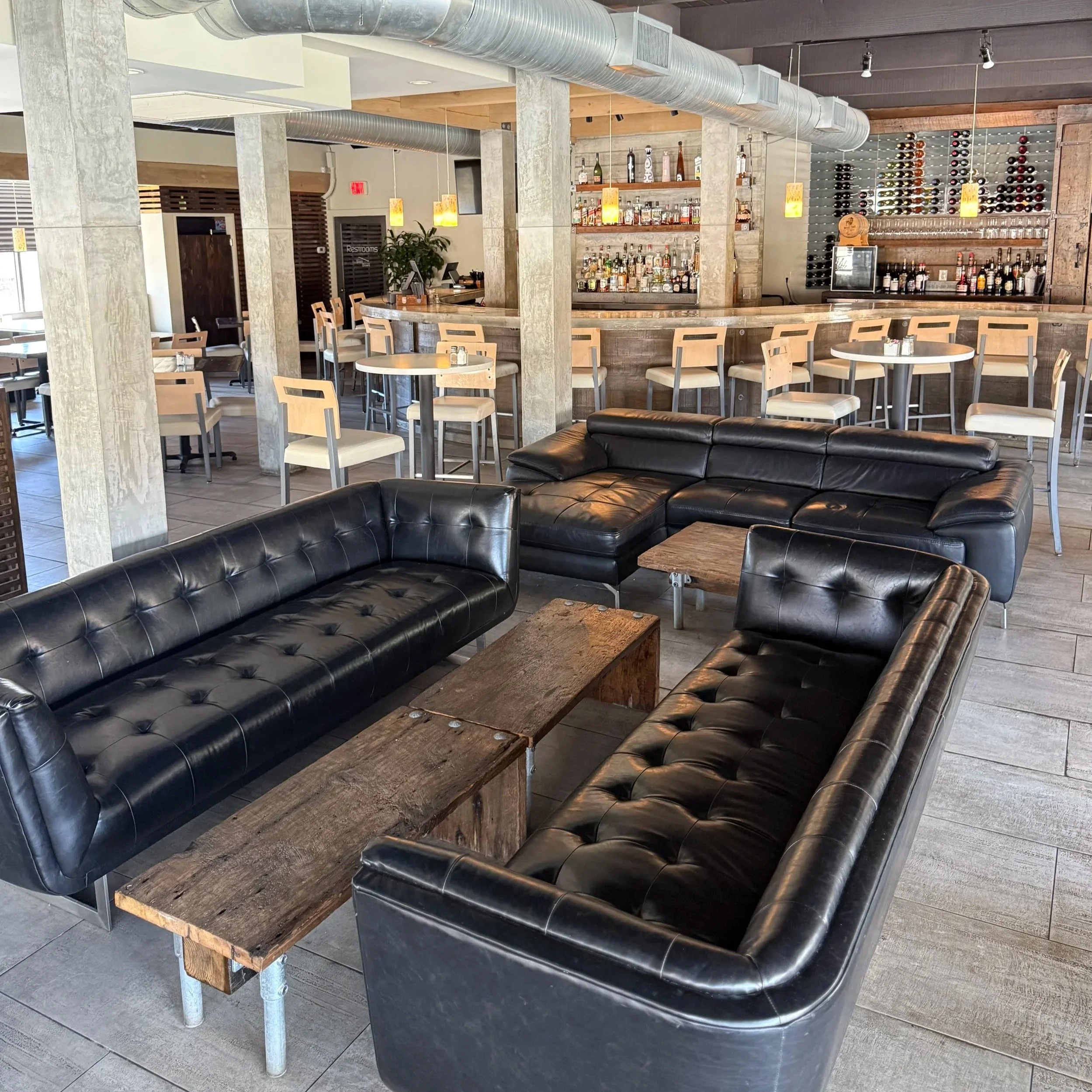 The image shows a bar area inside a restaurant with a wooden counter, liquor bottles, wine racks, and modern decor, including hanging yellow lights and a black leather sofa in the foreground.