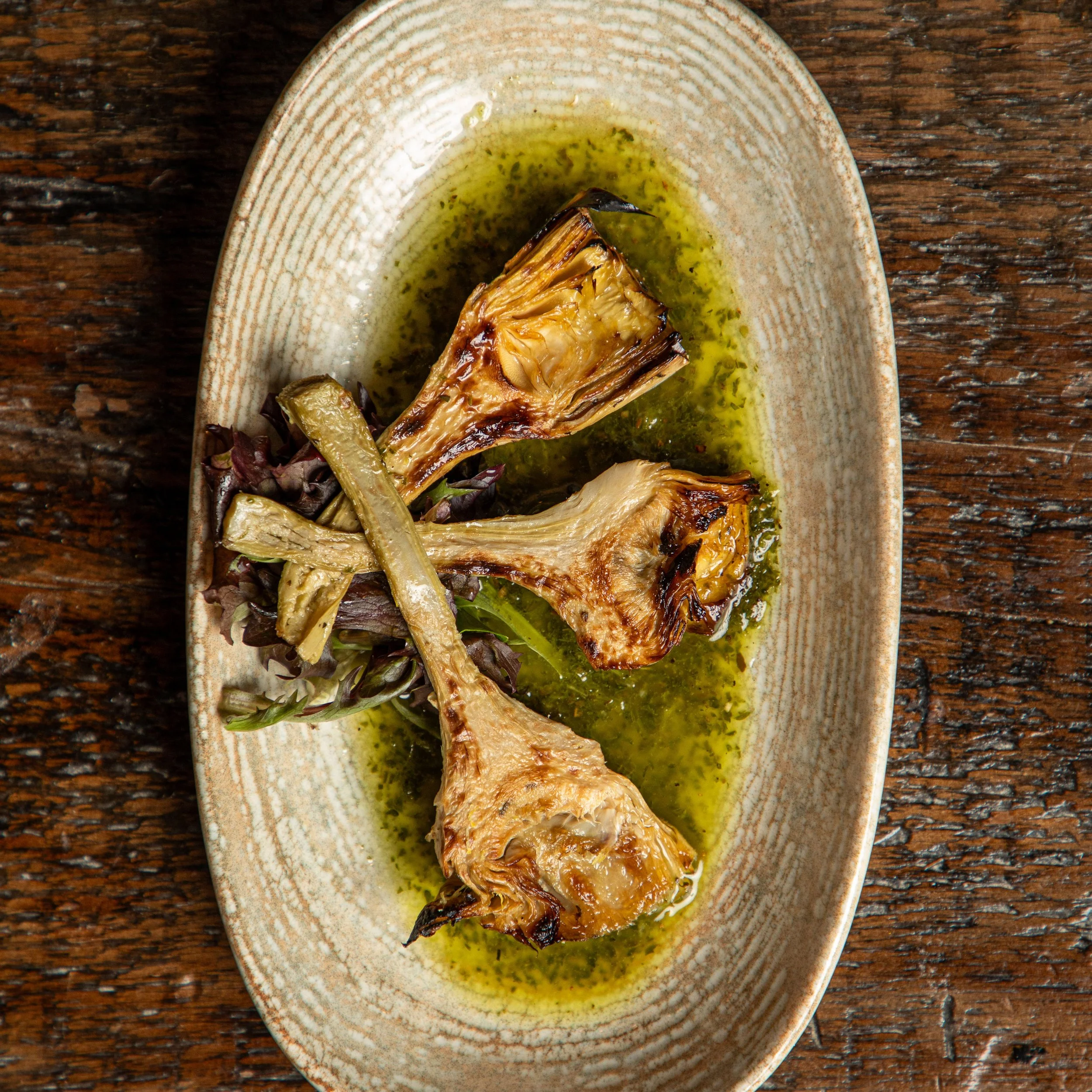 Three cooked lamb chops with herbs and sauce on a beige oval plate, garnished with greens, on a wooden table.
