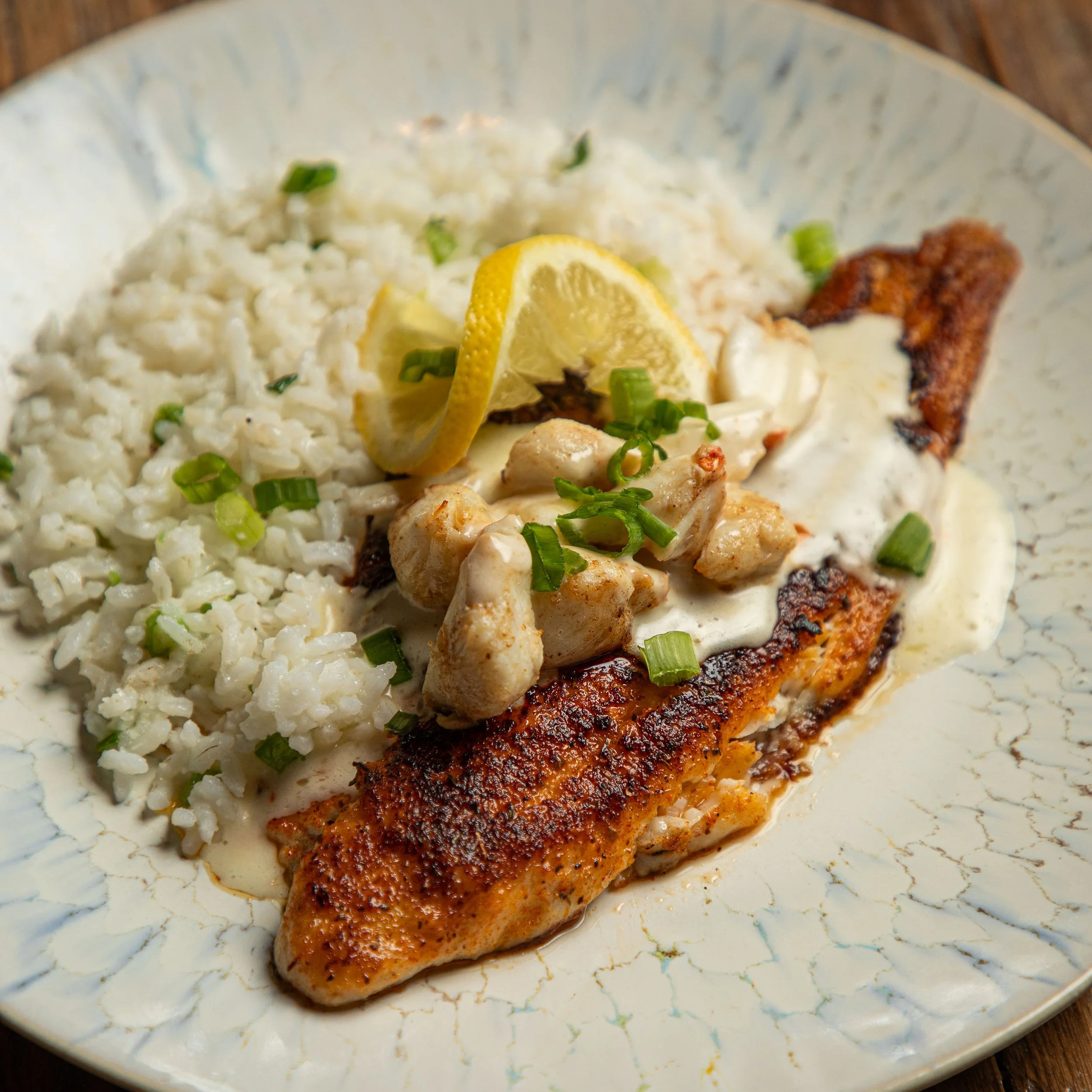 A plate of cooked fish with creamy sauce, served with white rice, garnished with lemon slices and chopped green onions.